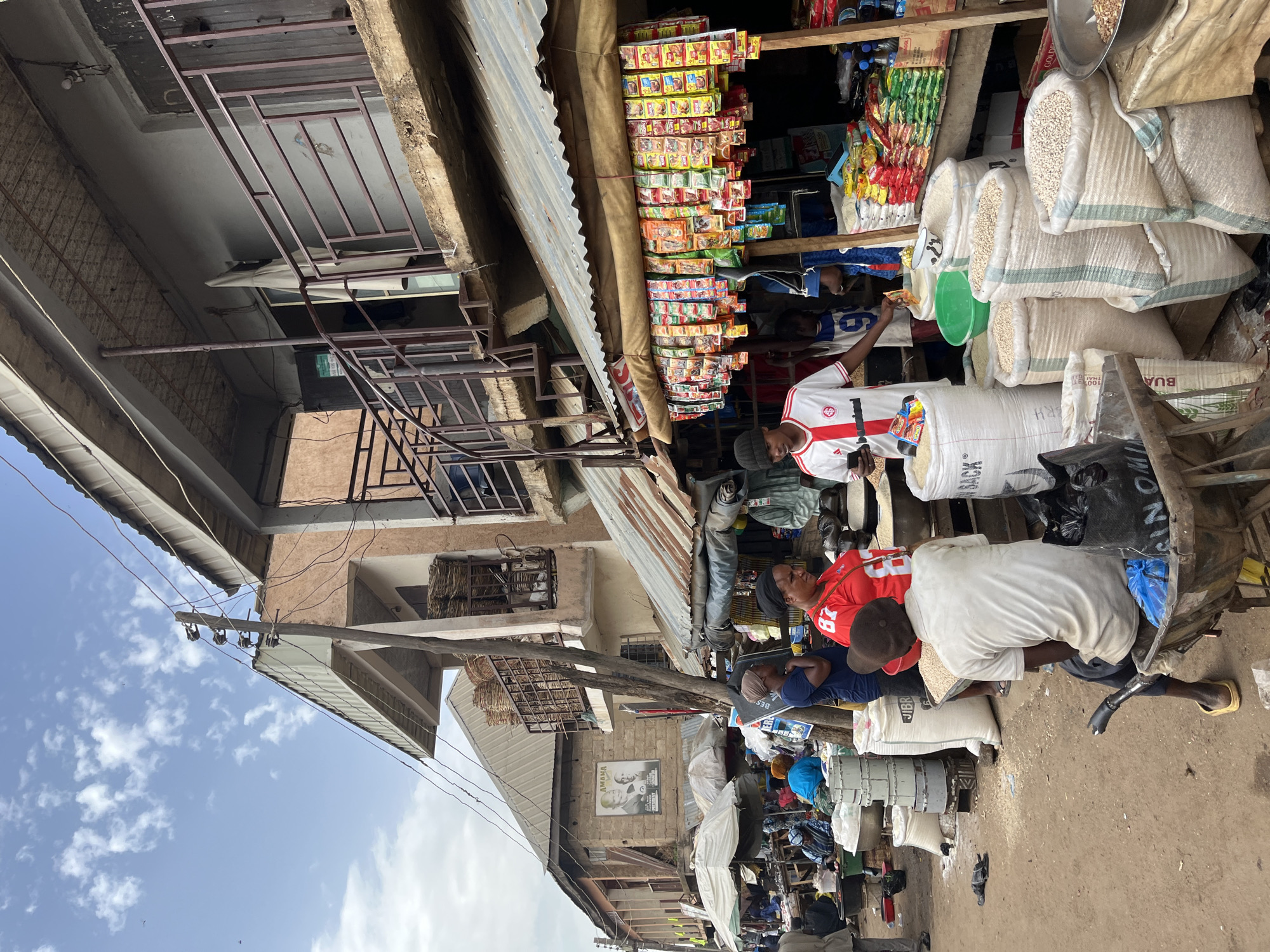 This image depicts a bustling outdoor market scene in Rafin Par, Nigeria, under a partially cloudy sky. The foreground is dominated by sacks of grain, primarily white and light-colored, some of which are partially filled. A man wearing a white t-shirt with red accents and a black beanie is seated on a wheelbarrow filled with grain, holding a mobile phone. Next to him, a woman in a red t-shirt with the number "87" on it stands and looks away from the camera. Another man, wearing a white t-shirt and dark pants, is in the background near a stall displaying numerous colorful packets of goods, possibly food items or spices, hanging from a corrugated metal awning.
The market appears to be organized along a street or pathway lined with structures. One building, situated behind the market stalls, features multiple levels with balconies and windows. A large poster with two men's faces is visible on the side of another building. In the background, more market stalls with umbrellas and various goods are visible, suggesting a diverse array of vendors and products. People are seen engaged in various activities, including shopping and attending to their stalls. The ground is dirt, and there are scattered items such as bags, containers, and what appear to be electrical wires overhead, typical of an active marketplace environment.