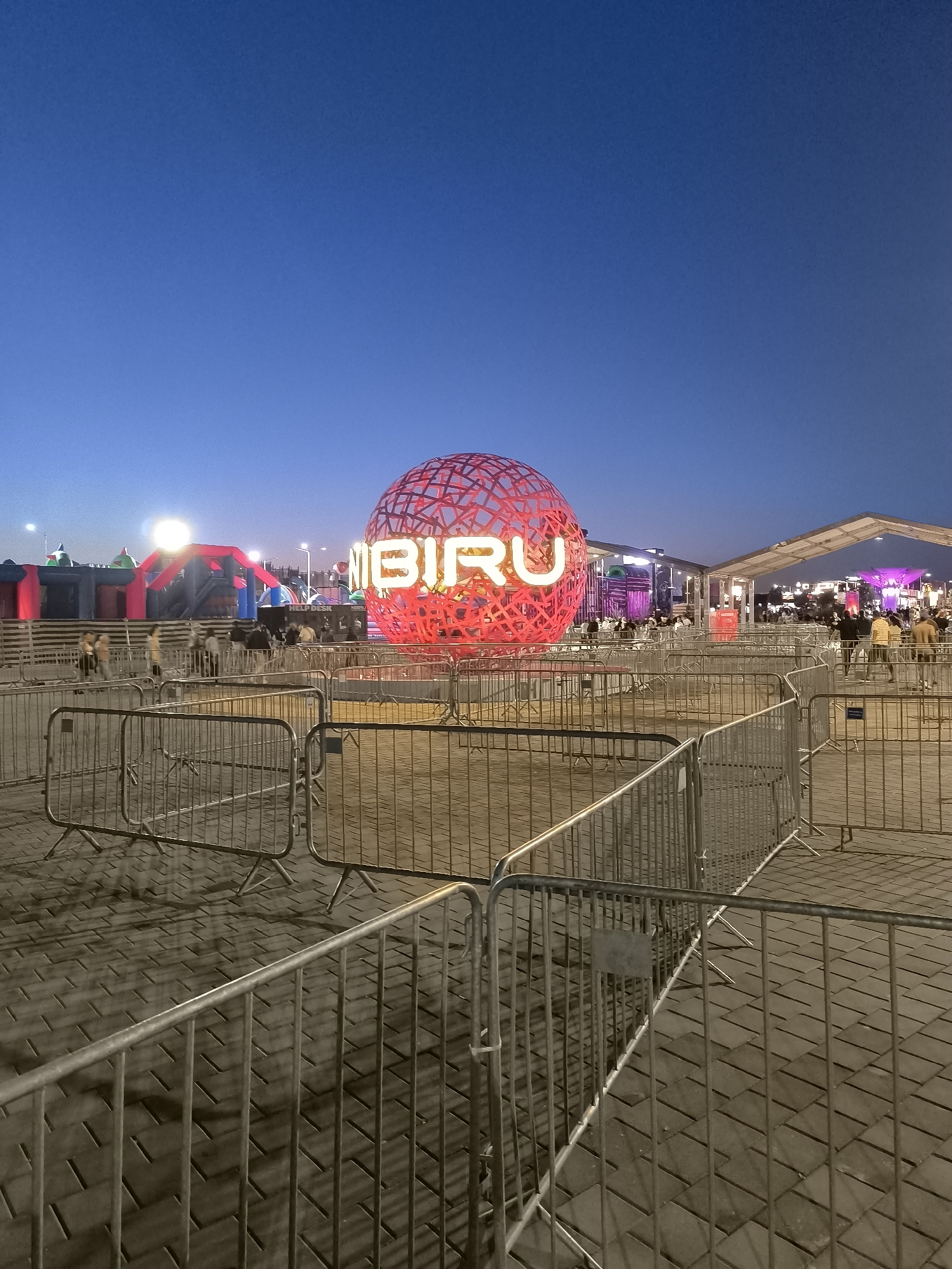 The image depicts an outdoor scene at dusk in Costinești, Romania, likely at an event or festival. In the foreground, several sections of silver metal crowd control barriers are arranged on a paved walkway. Behind the barriers, a large, illuminated red sphere with a lattice-like structure displays the word "NIBIRU" in glowing white neon letters. Further in the background, there are various structures, including what appear to be inflatable attractions or stages, tents, and groups of people. The sky is a deep blue, indicating the transition from daylight to night. The overall atmosphere suggests a lively, public gathering.