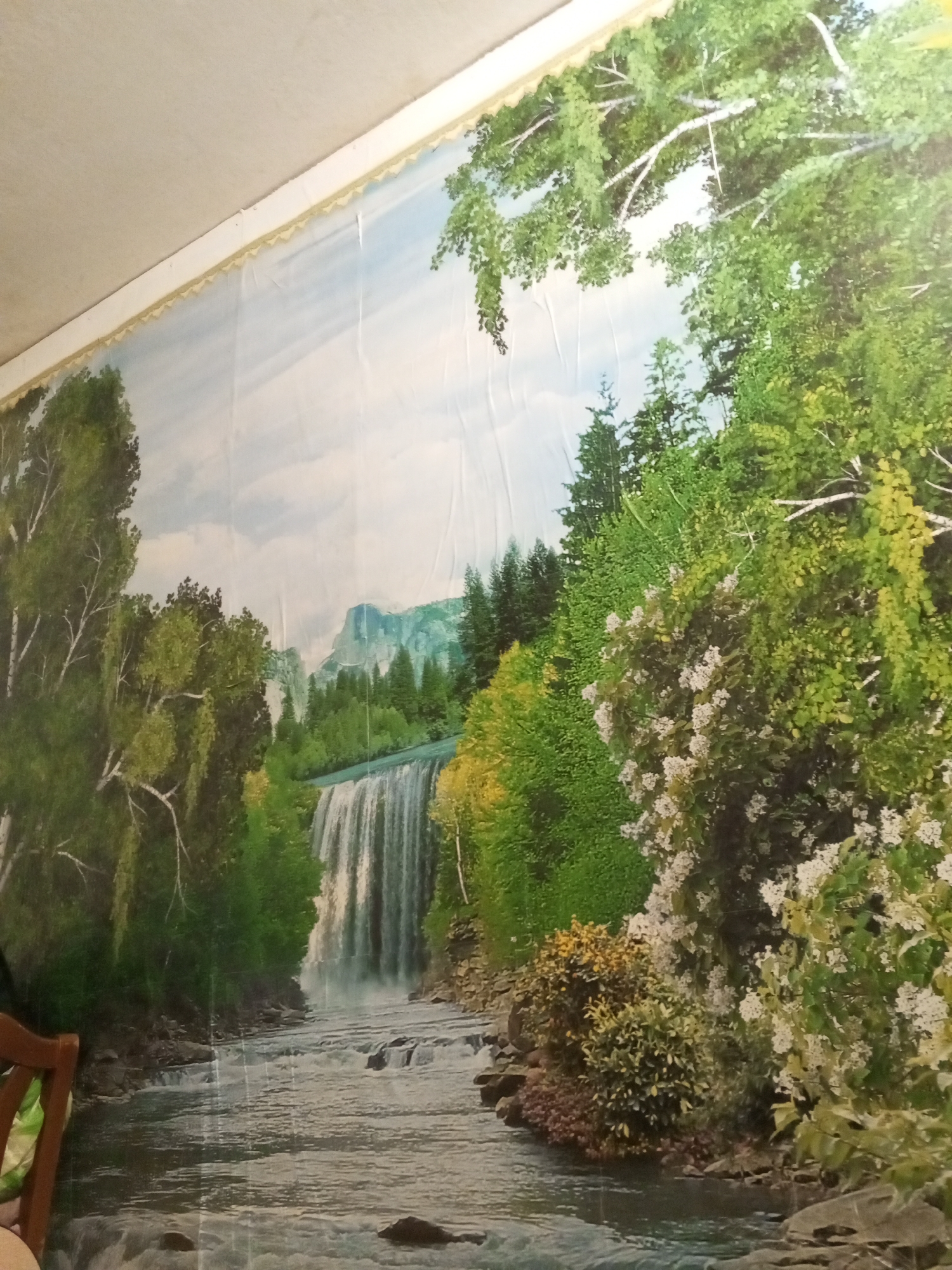 The image is a photograph of a mural depicting a natural landscape with a waterfall and a river. The mural covers a wall and extends upwards to meet the ceiling. The scene features lush green trees, a cascading waterfall, a rocky riverbed, and mountains in the distance. The sky is a soft blue with scattered clouds. A decorative trim runs along the top edge of the mural, just below the ceiling. There are no people or animals visible in the mural. The scene is static and painted, therefore no actions or interactions are taking place. The location in the city of Старое Ромашкино, Russia, is not visually evident from the mural itself, but the photographer has specified it.