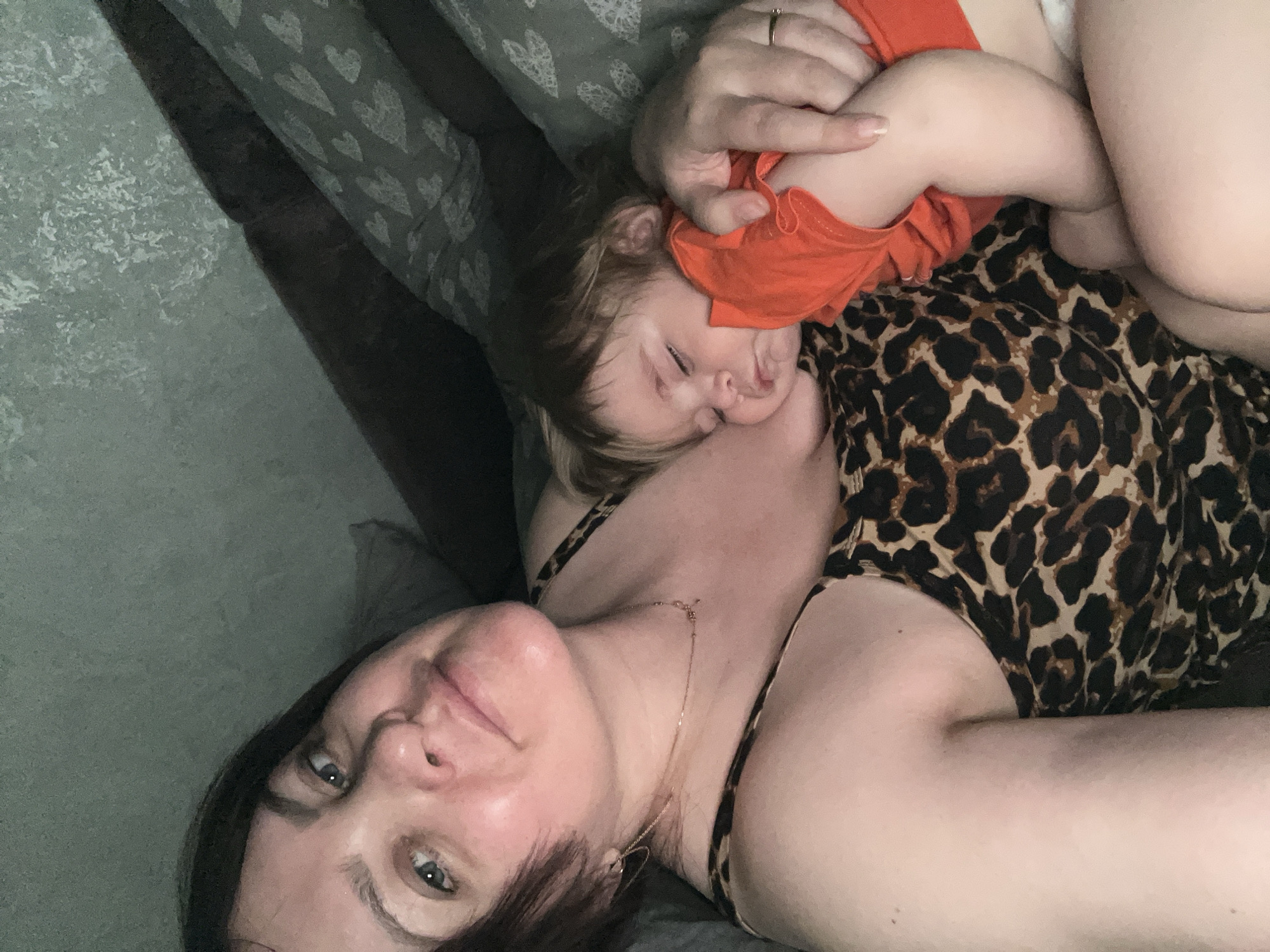 A medium, close-up shot shows a woman lying on her back with a sleeping baby nestled on her chest. The woman has fair skin, dark hair, and blue eyes. She is wearing a cheetah-print top with thin straps and a gold necklace with a heart-shaped pendant. The baby, dressed in an orange shirt, is lying face down against her collarbone with closed eyes. One of the woman's hands is gently resting on the baby. They are on a gray surface with a heart-patterned pillow visible in the background. There is no evidence this image was taken in the city of Noviy, Russia.