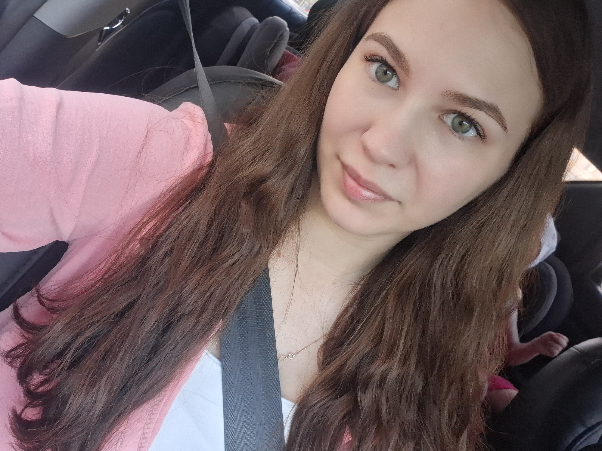 A woman with long brown hair is sitting in the driver's seat of a car, wearing a pink jacket over a white top. She is wearing a seatbelt and has green eyes. In the back seat, a child's hand is visible, suggesting a child is present. The car's interior is visible, with black seats and some baby/child car seats. The location of this image is unknown and cannot be determined using the location of Chaikovsky, Russia.