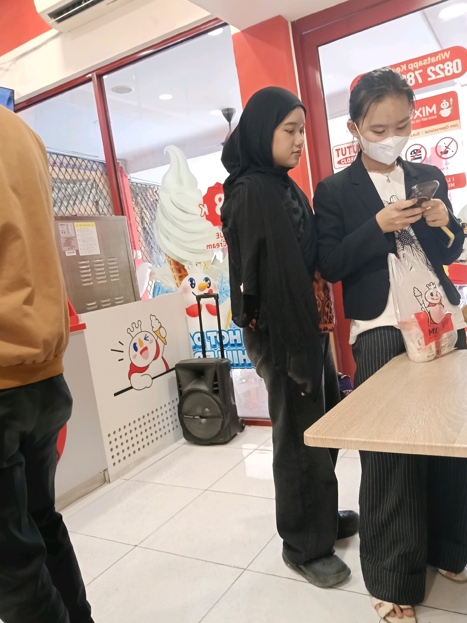The image shows two young women in what appears to be a food establishment in the city of Belimbing, Indonesia. The woman on the left is wearing a black hijab and a black outfit. She is looking to her left. The woman on the right is wearing a black blazer over a white t-shirt and pinstripe pants. She is wearing a white face mask and looking down at her mobile phone, which she is holding in her hands. She is also holding a plastic bag with a logo on it. In the background, there is a large inflatable ice cream cone, a speaker, and signage that appears to be in Indonesian. The lighting is bright, suggesting an indoor setting. The floor is tiled.