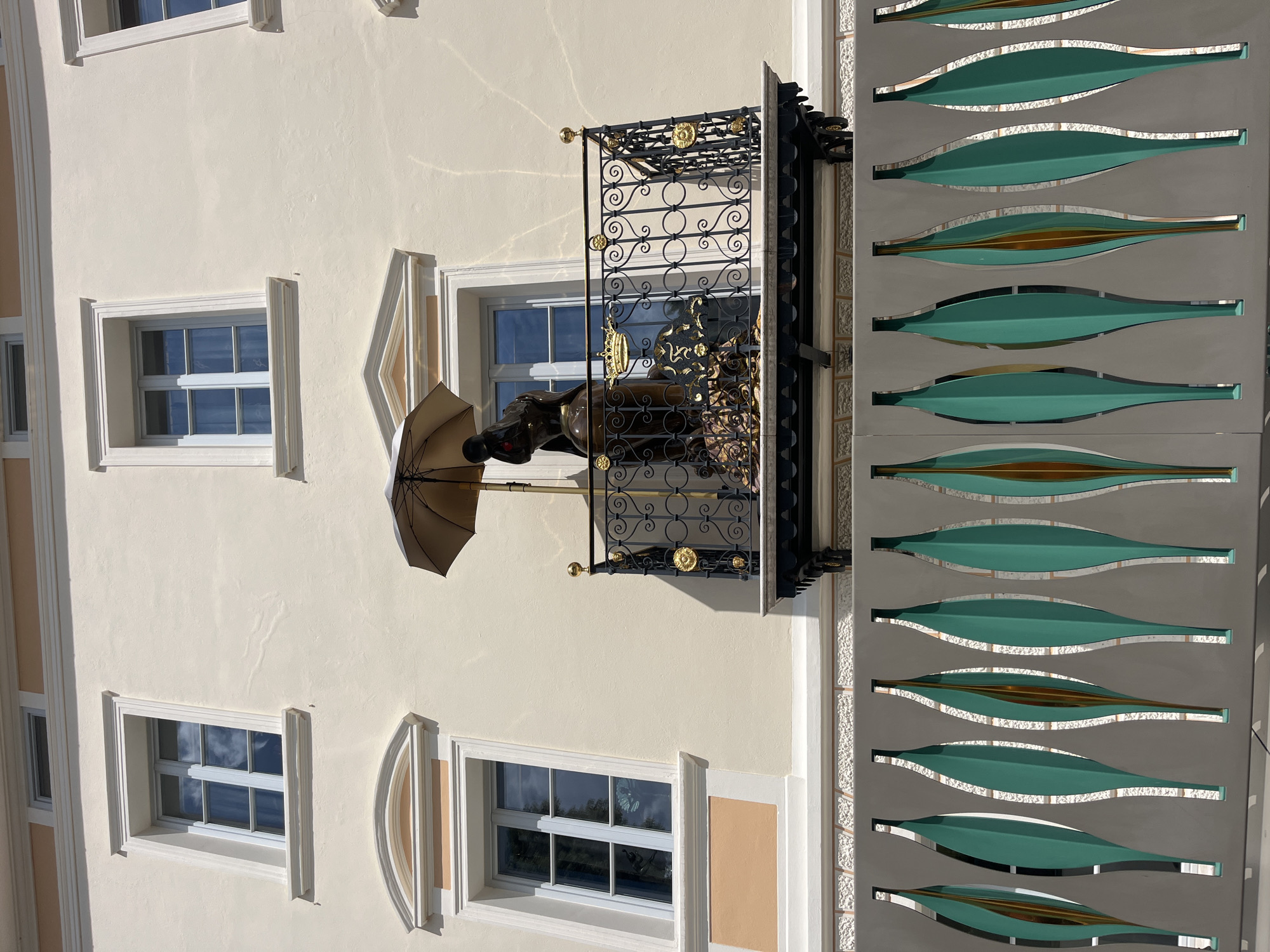 The image depicts the facade of a building in Brixen - Bressanone, Italy, featuring a balcony with a bronze sculpture. The building has a light-colored stucco exterior with cream-colored window frames and decorative peach-colored accents. There are three visible windows on the upper floor, two of which are partially obscured by the building's architecture. The balcony, made of black wrought iron with gold ornamentation, holds a bronze sculpture of a seated figure holding an umbrella. The sculpture is detailed, showing the figure's posture and facial features, and appears to be adorned with a crown. Below the balcony, a decorative fence with teal and gold leaf-shaped panels is visible, creating a layered effect with the building. The lighting suggests a bright, sunny day, with shadows cast on the facade.