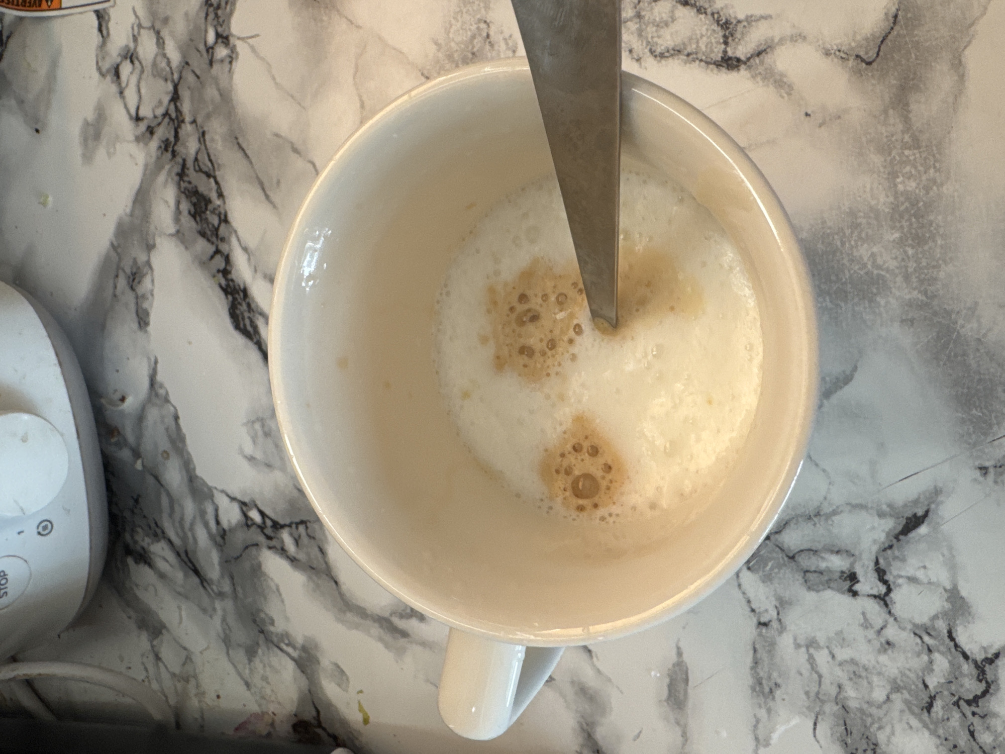 This is a top-down, close-up shot of a white ceramic mug filled with a frothy, light brown beverage, likely coffee or a similar drink. A silver spoon is partially submerged in the liquid, indicating that the drink is being stirred or has just been prepared. The foam on the surface of the drink has small bubbles and some darker, concentrated areas, suggesting the presence of espresso or coffee.
The mug is placed on a countertop with a marble-like pattern, featuring veins of black and grey against a white background. To the left of the mug, a portion of a white electronic device is visible. It has a circular dial and buttons, one labeled "STOP" and another with a timer icon. This device appears to be a kitchen appliance.
The lighting suggests it could be daytime, possibly indoors in a kitchen or dining area. There are no explicit indicators of the weather or specific time of day beyond general daylight. The scene is clean and tidy, with no visible mess. The overall mood is calm and domestic, possibly capturing a moment of preparation for a beverage.
There is some visible text on a sticker partially visible at the top left of the image, but it is mostly obscured and illegible. The text on the appliance buttons is clear: "STOP" and a circular symbol that appears to be a timer or clock.