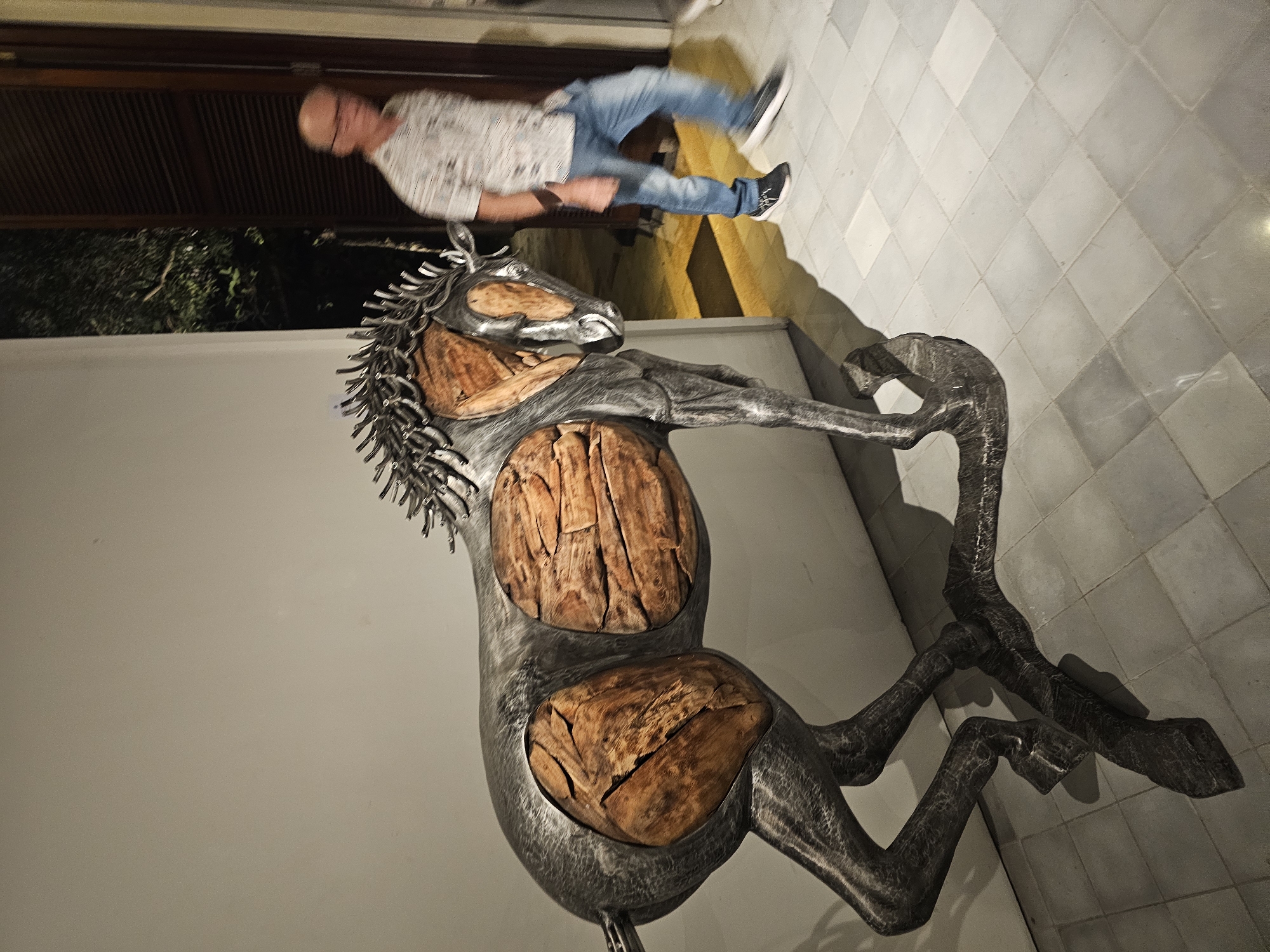 A full shot captures a metallic horse sculpture with wooden inlays, positioned near a white wall and a tiled floor. A person is seen above, wearing a light-colored shirt and blue jeans, with his feet in black shoes. The scene may be located in Denpasar, Indonesia.