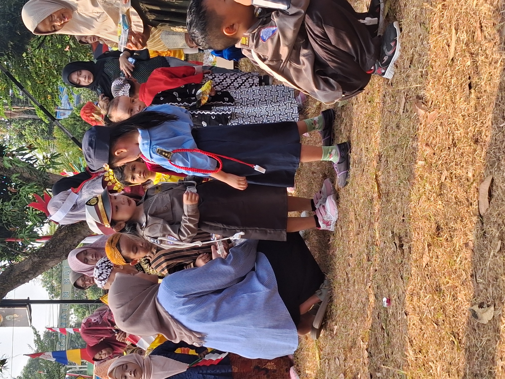 The image depicts an outdoor scene in Balapulang, Indonesia, possibly during a local event or celebration, given the presence of children in uniforms and adults observing. The ground is covered with dry grass and fallen leaves, indicating a warm, possibly dry climate.
In the foreground, a young girl in a blue shirt, dark skirt, and dark cap stands facing left, her right arm extended as if speaking or gesturing. A red cord with a white pendant hangs from her neck. To her right, a young boy in a brown police uniform with patches on the sleeves and chest is crouched down, looking towards the right. His left hand is near his mouth.
Behind them, other children and adults are visible. A woman wearing a light brown hijab and a patterned dress is holding a small, clear plastic container with food or candy. She is looking down and speaking to someone off-camera. Another woman, partially obscured, wears a light blue striped shirt and a dark skirt, and is crouched down. Several other children are dressed in various colorful attire, some in traditional Indonesian patterns. Indonesian flags and banners are visible in the background, suggesting a festive or patriotic occasion. The overall atmosphere appears to be one of community gathering and activity.