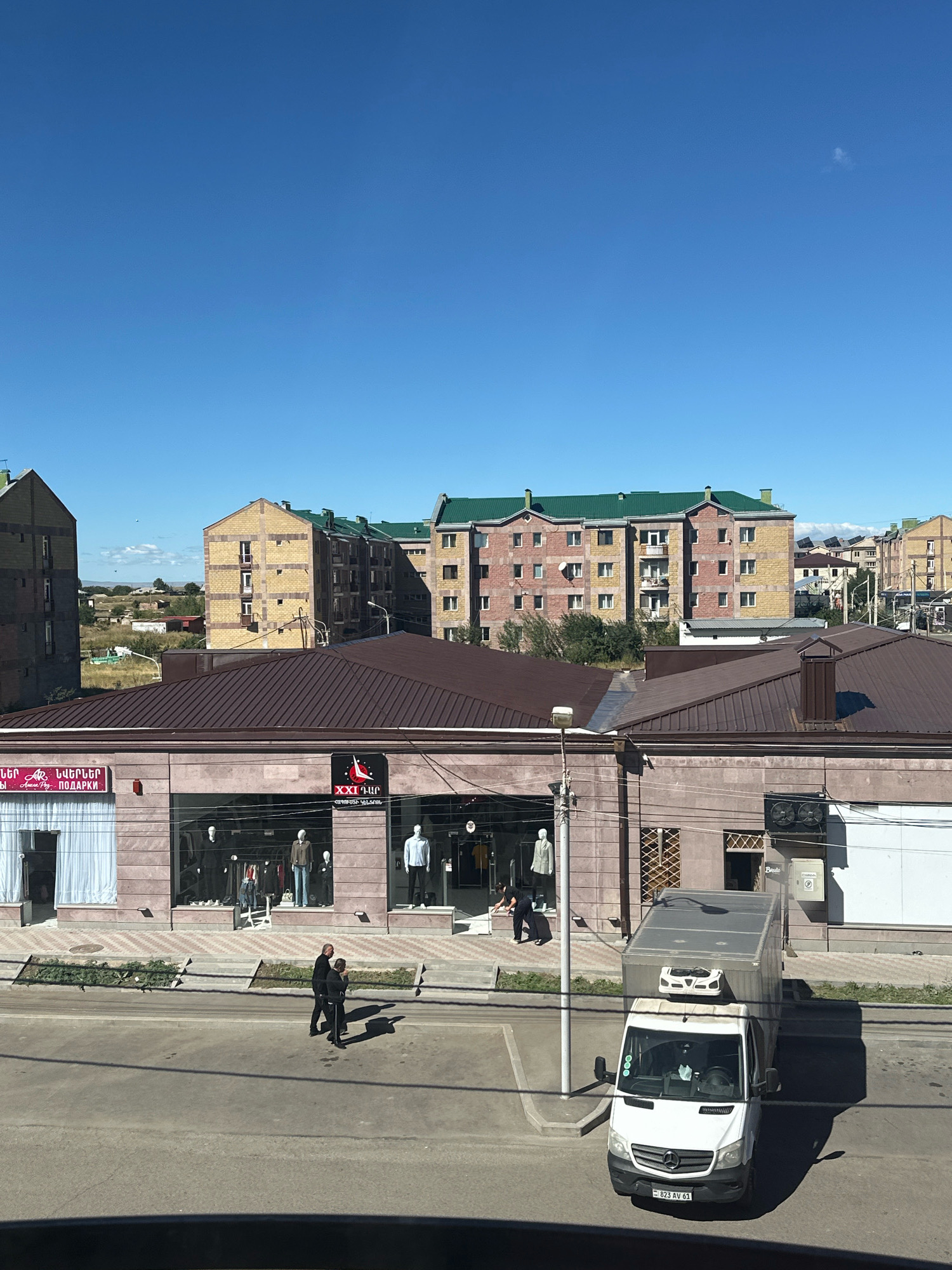 This is an exterior, high-angle shot of a street scene in Gyumri, Armenia, on a clear, sunny day with a bright blue sky. In the foreground, a white refrigerated truck is parked on the right side of the street. Two people, a man and a woman, are walking across the street from left to right in front of a commercial building. The building has a brown, metal-clad roof and a facade with large display windows. Inside the display windows, mannequins are dressed in clothing. Above the display windows, there are signs with text in Armenian. In the background, there are multi-story residential buildings with light-colored facades and green roofs. The street is paved, and there is a sidewalk with some greenery in planters. The overall scene depicts a typical urban environment with commercial and residential areas.