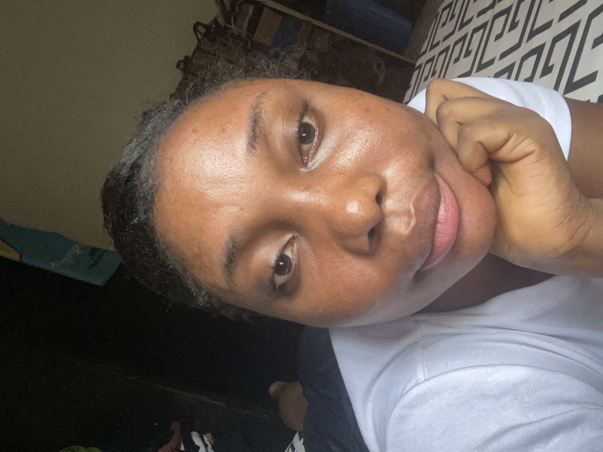 A person is lying down with their head resting on their hand, looking directly at the camera. They have dark hair and brown skin. They are wearing a white t-shirt. The background includes a patterned bedspread and a dark-colored surface. There's no information to suggest this image was taken in Abuja, Nigeria.