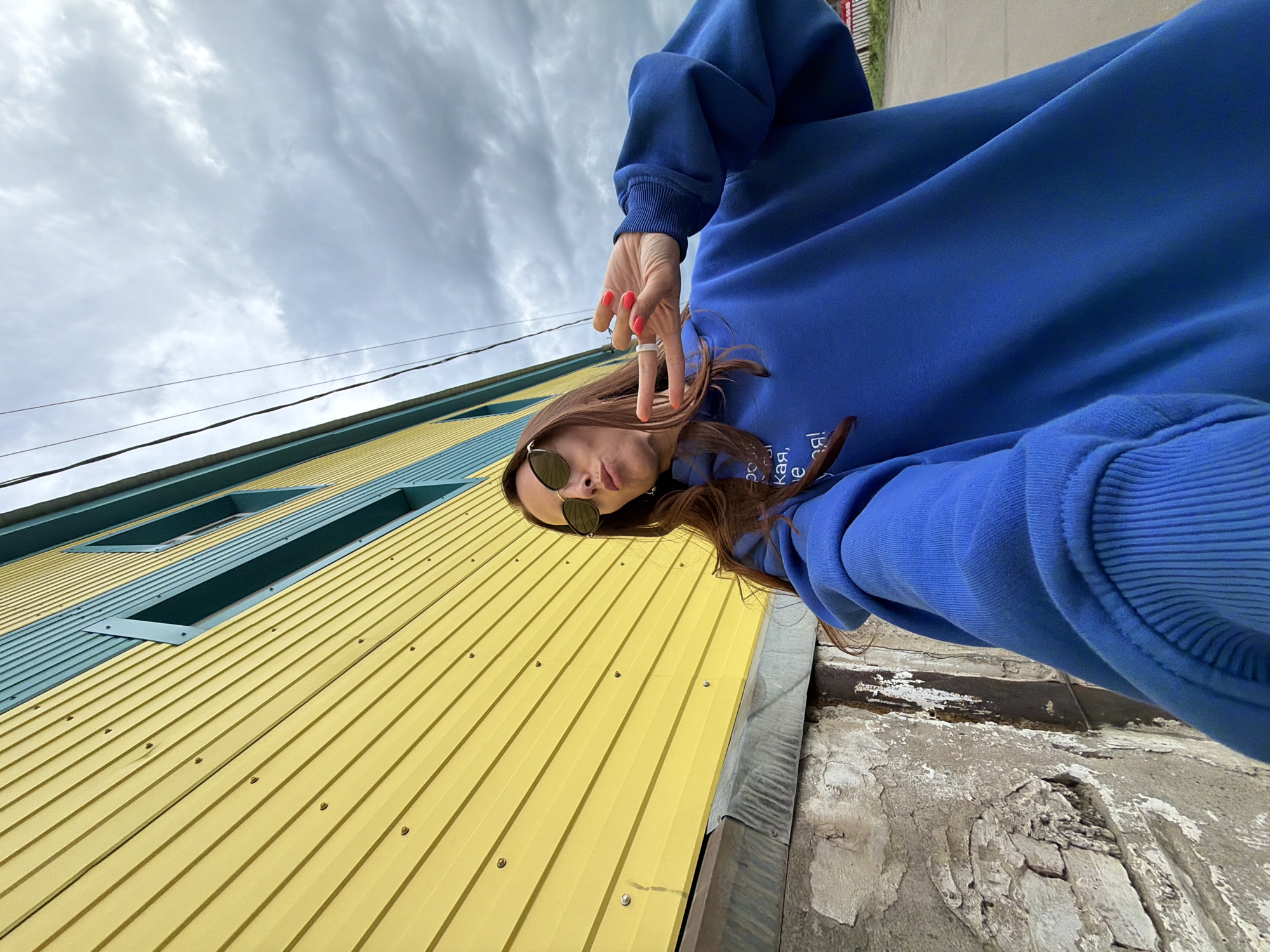 This low-angle, upside-down selfie captures a young woman against a vibrant building facade and an overcast sky. The subject, positioned in the upper center of the frame, has long brown hair and wears dark sunglasses and an oversized royal blue sweatshirt or hoodie. Her right arm is extended towards the viewer, with her hand making a peace sign. She has coral-colored nail polish and a prominent white ring on her middle finger. Partially visible Cyrillic text on her sweatshirt reads "ая, ве я!".
The background features a building clad in bright yellow corrugated metal siding. A horizontal strip of teal or turquoise corrugated metal runs across the upper section of the building, containing several rectangular openings that appear to be windows or vents. Above the building, the sky is overcast with soft, diffused clouds, and two dark power lines stretch horizontally across the upper part of the image. The immediate foreground to the bottom right shows a weathered and textured surface of concrete or stucco with peeling paint, adjacent to a metal downspout or gutter. The scene appears to be set outdoors during the daytime under cloudy conditions, in Birobidzhan, Russia. The unusual perspective suggests a creative or playful approach to photography.
