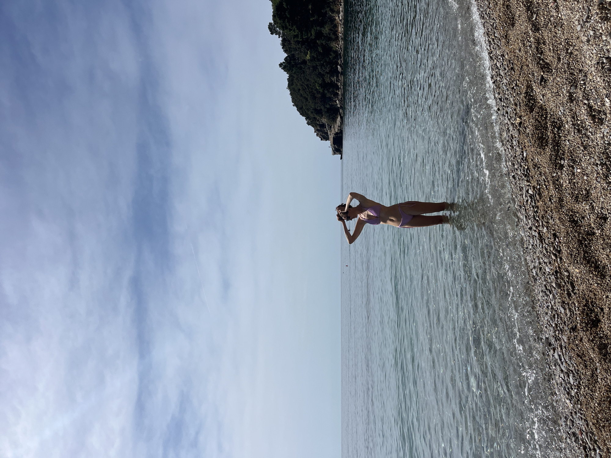 A vertical, long shot shows a woman in a pink swimsuit standing in shallow water in Budva, Montenegro. The woman is centered and faces right, with her arms raised near her head. To the right of the frame, a rocky beach transitions into clear, shallow water. The water surface has gentle ripples. In the distance, a tree-covered landmass appears on the upper right of the frame. The sky is light blue, with thin, streaky clouds on the left.