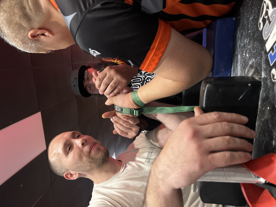 Three men are arm wrestling. One man, wearing a white shirt, is seated at the table with his arm secured by a green strap. Another man in a black and orange shirt is next to him. A third man, wearing a baseball cap and glasses, is watching them. The arm wrestling table is black and white.