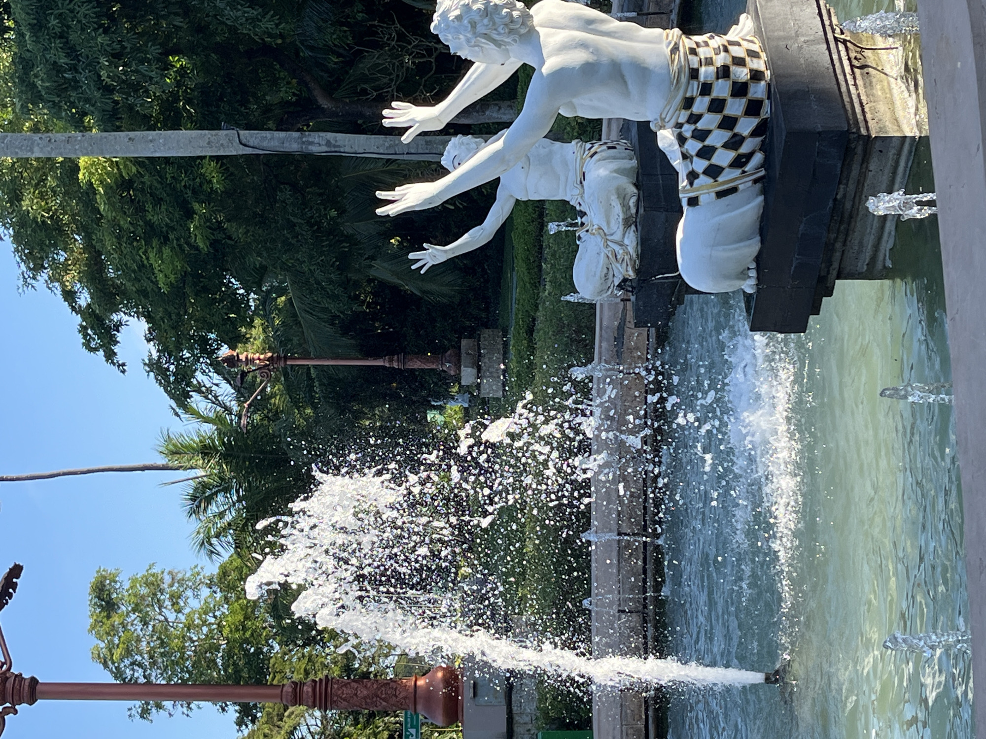 The image shows a fountain featuring two kneeling, white-painted statues of human figures with outstretched arms, likely male. The figures wear garments or sashes with a black and white checkered pattern. Water spouts from multiple locations within the fountain, including a central jet that creates a spray. The fountain is surrounded by greenery, including trees and shrubs, suggesting an outdoor setting. A lamppost with a decorative design is visible on the left side of the frame. The location is in the city of Unknown, Indonesia.
