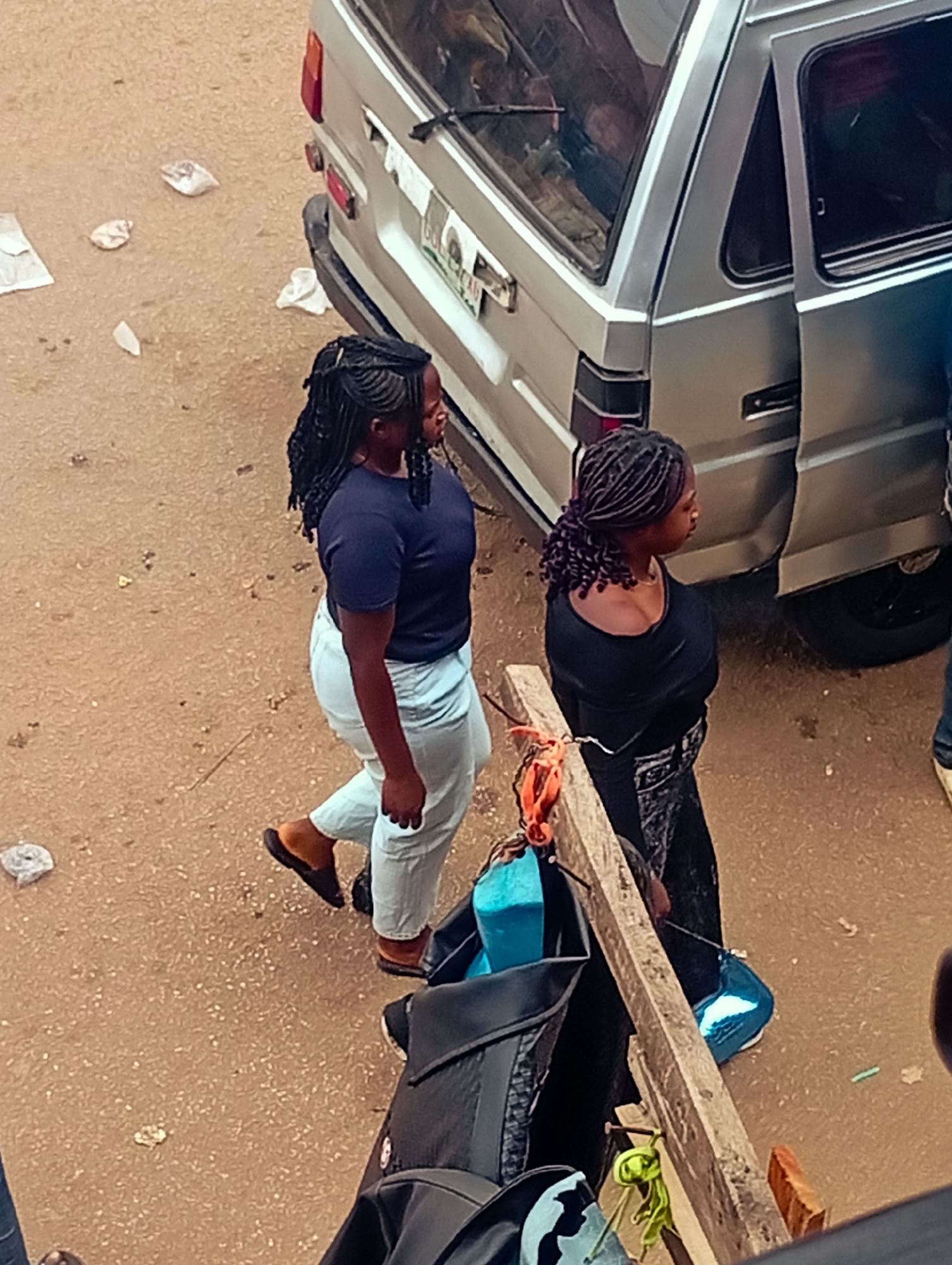 Two Black women with braided hairstyles are walking past a silver van on a dirt street in a city of Unknown, Unknown. The woman on the left is wearing a dark blue top and light blue jeans, while the woman on the right is wearing a black top and a black and white patterned skirt. A wooden structure with bags and other items is in the foreground. There are pieces of trash scattered on the ground. The van's rear door is open, and a person is standing near it.