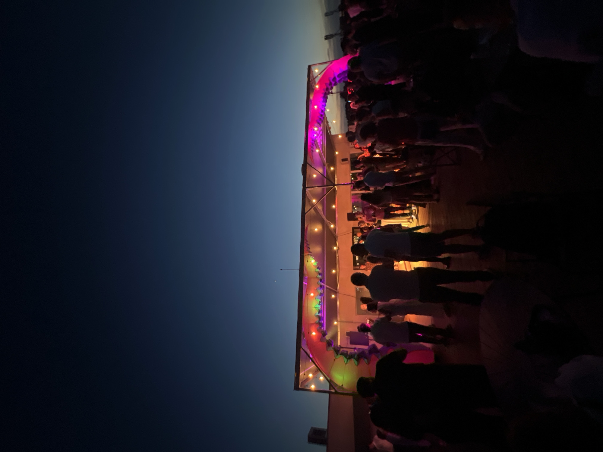 A rooftop party is in full swing at dusk, with a clear, dark blue sky overhead. A structure, possibly a stage or bar, is illuminated by colorful lights in pink, green, and purple hues, casting a vibrant glow on the scene. String lights are also visible, adding to the festive atmosphere. A crowd of people, mostly silhouetted against the lights, are gathered on the rooftop, some standing and observing, while others appear to be interacting or dancing. The foreground is largely dark, obscuring many details of the people present. The overall impression is a lively evening social event. The specific location in Sofia, Bulgaria, cannot be determined from the visual information provided.