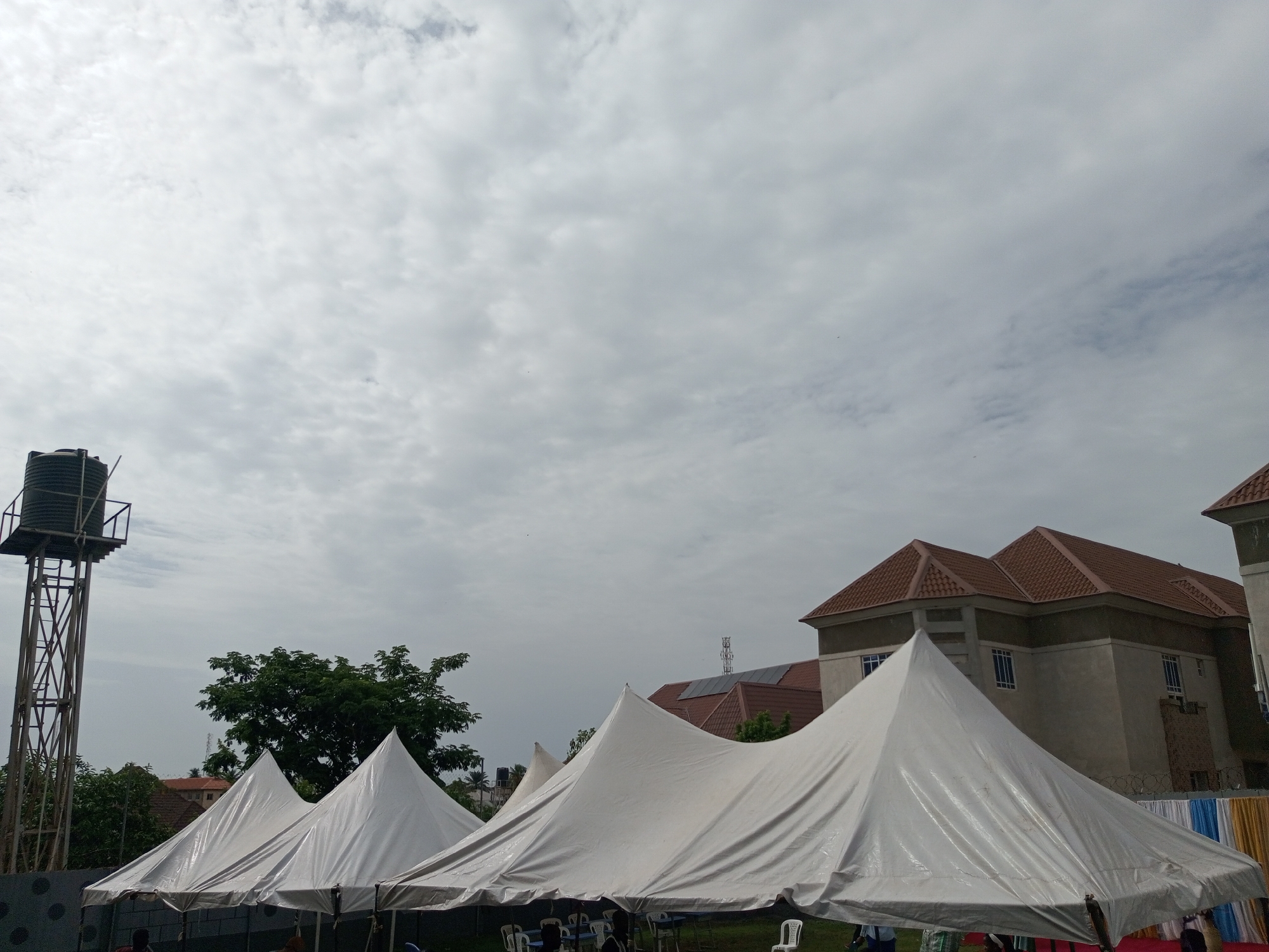 The image depicts an outdoor scene under a cloudy sky, likely in Barden Yana, Nigeria, given the architectural style and vegetation. In the foreground, several white event tents are set up. The tents have pointed peaks and are made of a reflective material. Some tables and chairs are visible beneath the tents, suggesting an event is being prepared or is underway.
To the left, a tall metal structure supports a large water tank, a common sight in areas with water supply challenges. Behind the tents and to the right, a multi-story building with a reddish-brown tiled roof is prominent. The building has multiple windows with white frames. Further in the background, more buildings and lush green trees are visible. A decorative fabric display, featuring colorful bands of yellow, blue, and white, is visible along a fence in the middle right of the image. A few people are partially visible under the tents, engaged in what appears to be setting up or attending the event. The overall atmosphere is that of an outdoor gathering or celebration.
