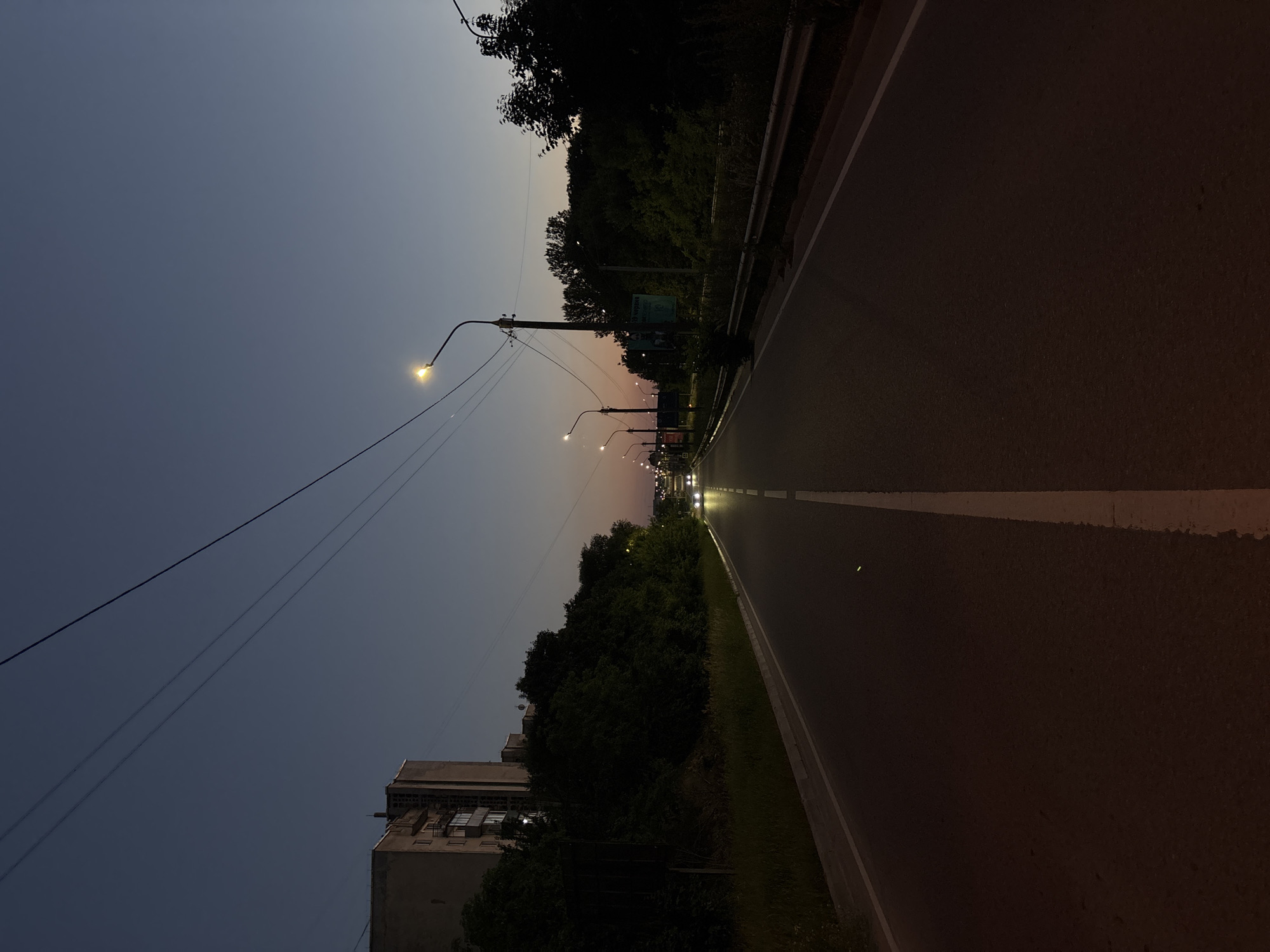 The image shows a dimly lit road at dusk or dawn. The sky is a gradient of deep blue to a soft pinkish hue near the horizon. Several streetlights are illuminated, casting a warm glow on the road and the surrounding trees. The road itself appears to be asphalt and has a white line marking the lane.
On the left side of the road, there is a dense line of trees, and beyond them, a multi-story building is partially visible. The building is light-colored and appears somewhat aged, with visible windows and architectural details. Several power lines are strung across the sky, connecting to poles that also support the streetlights.
In the distance, more streetlights and the headlights of vehicles can be seen, suggesting the road is a route with some traffic. A metal guardrail runs along the edge of the road, separating it from the vegetated area. The overall atmosphere is quiet and serene, with a hint of the transition from day to night or night to day. There are no people visible in the image. No discernible text is visible.