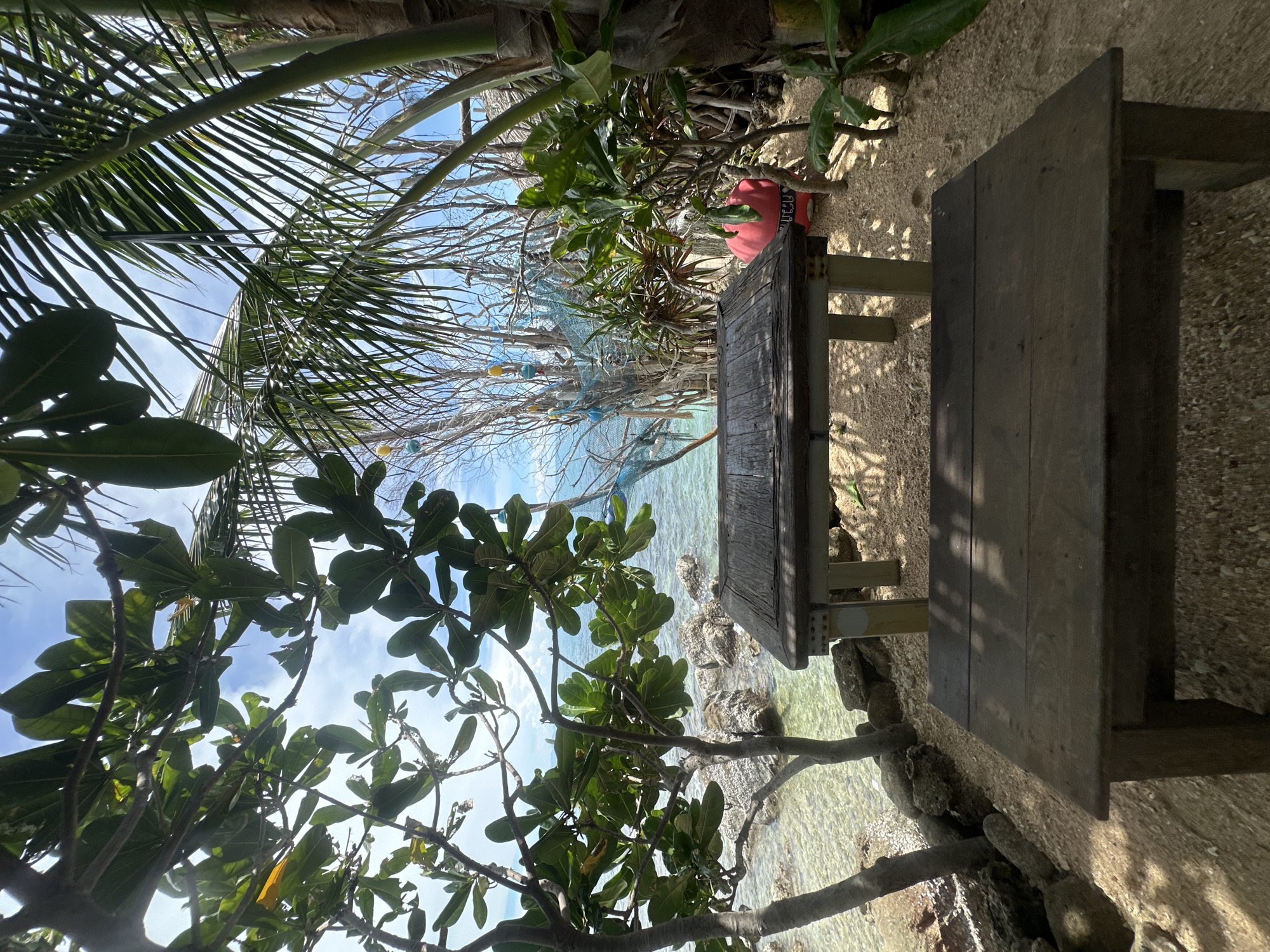 The image depicts an outdoor scene with a focus on a wooden picnic table and bench situated on a sandy area near the ocean. Tropical vegetation, including palm fronds and leafy branches, frames the view, suggesting a warm, sunny environment. The clear, shallow water of the sea is visible, with rocks and a shoreline in the foreground. In the background, a blue fishing net is strung between trees, adorned with colorful buoys. A bright red float with some text on it is also visible near the table. The overall impression is one of a tranquil, tropical paradise, likely a beach or seaside resort. The time of day appears to be daytime, with sunlight casting shadows. No people are visible in the image. The location context indicates Koh Phangan, Thailand.