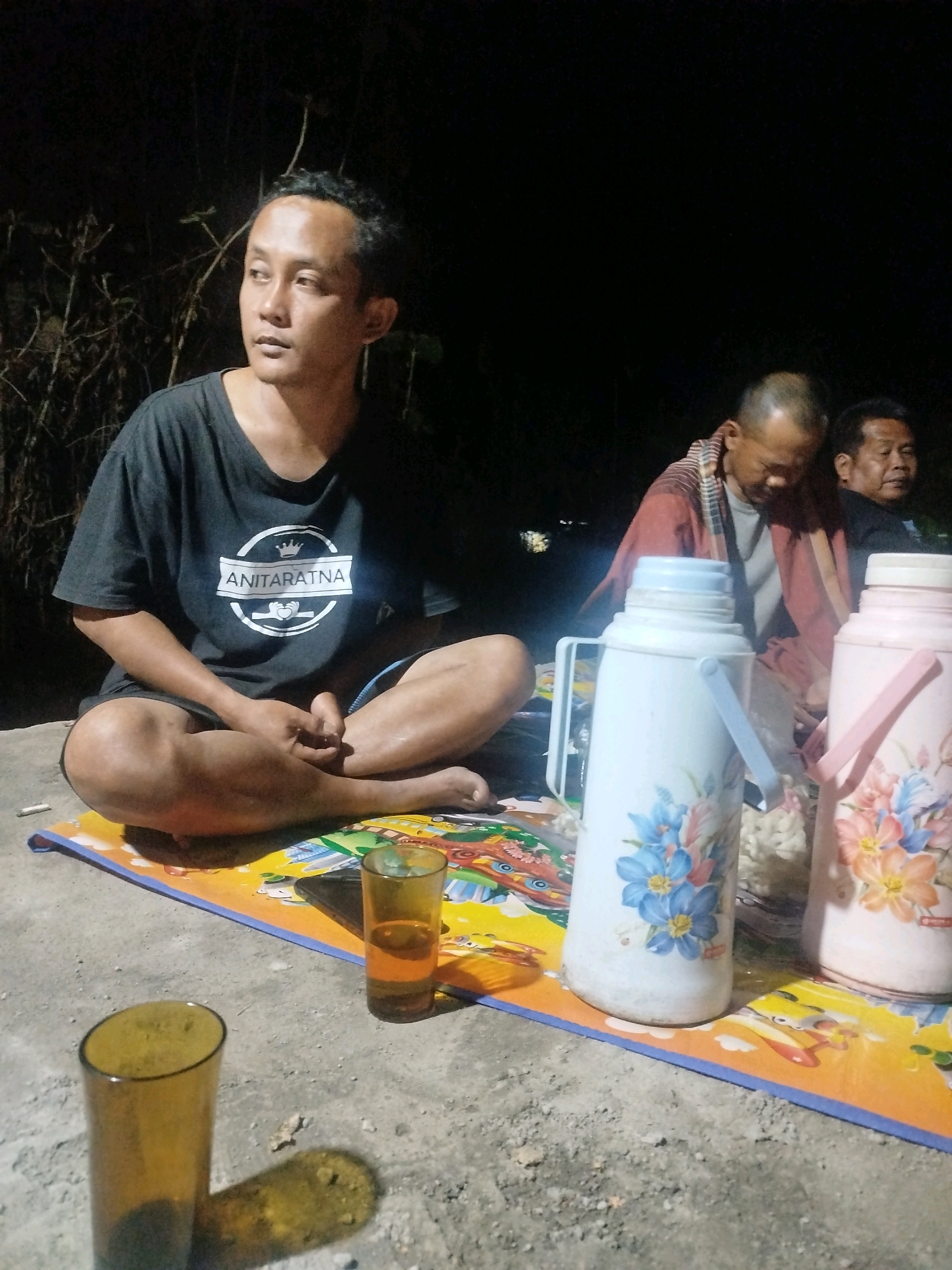 In the city of Semin, Indonesia, three individuals are gathered outdoors at night. The primary subject, a man wearing a black t-shirt with a white logo reading "ANITARATNA," is seated cross-legged on a colorful mat on the ground. To his right, partially obscured by a large white thermos decorated with blue flowers, another man is visible, leaning forward with his head down, wearing a reddish-brown patterned garment over a gray shirt. Further to the right, a third man is seated, looking to the left. In the foreground, two glasses are present; one is filled with an amber liquid, and another is empty. Two large thermoses, one white with blue flowers and a pink one with orange and blue flowers, stand on the mat. The background is dark, with some faint out-of-focus lights and foliage.