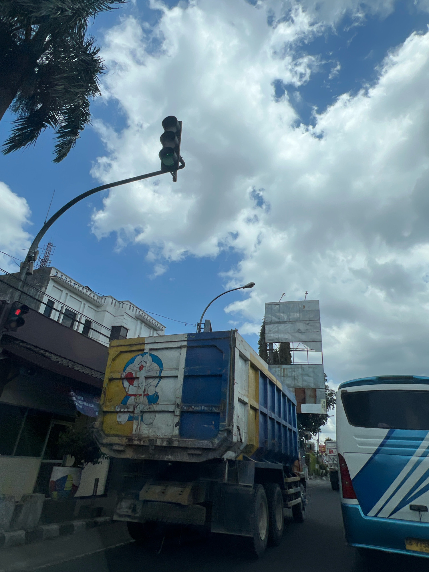 The image depicts a street scene in Magelang, Indonesia, under a partly cloudy sky. A large dump truck, adorned with a cartoon image of Doraemon on its side, is positioned in the foreground, blocking a significant portion of the view. To the right of the truck, the rear of a white and blue bus is visible. A traffic light, showing a red signal, hangs over the road, suspended from a black curved pole. In the background, several buildings line the street, with one white building featuring architectural details. A tall, metallic structure, possibly under construction or a billboard frame, stands further down the road. Palm fronds from a tree are visible in the upper left corner of the image. The overall scene suggests a busy urban environment with vehicles on the road.