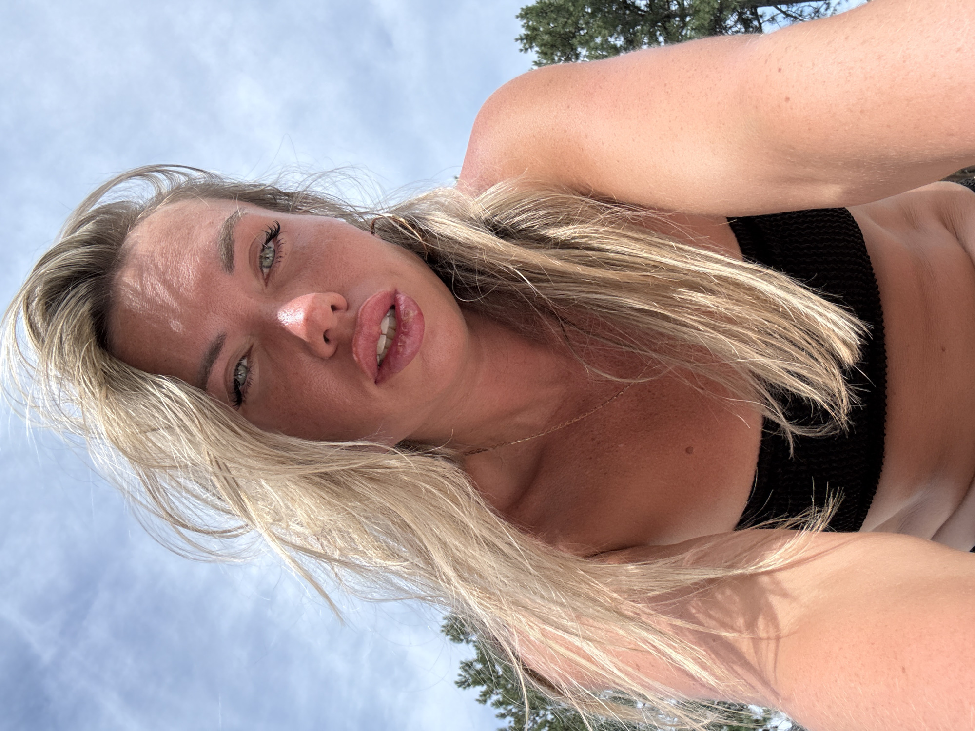 The image is a close-up, low-angle selfie of a blonde woman outdoors. She is wearing a black bikini top and has her head tilted back, with her face dominating the frame. Her eyes are open and looking towards the camera, and her lips are slightly parted, revealing her teeth. Her blonde hair is long and appears windswept. The background consists of a cloudy sky and the top of a green tree, suggesting it is daytime. The lighting indicates it is a sunny day. There is no visible text in the image.