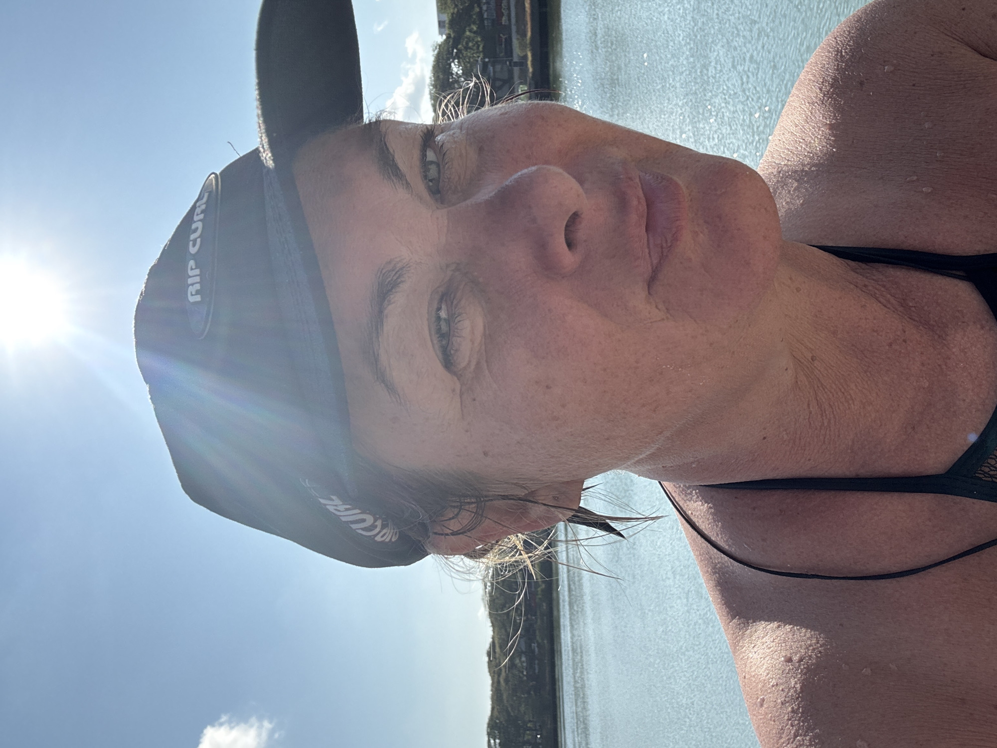 A close-up, angled selfie shows a woman wearing a black baseball cap with "Rip Curl" visible on the side. She has fair skin with freckles, light eyes, and damp blonde hair. Water droplets are visible on her face and chest. She is wearing a black swimsuit with thin straps. The background features water, possibly a lake or bay, with shoreline vegetation visible in the distance. The bright sun is in the upper left corner of the frame, causing a lens flare. The specific location in Baan Chaweng, Thailand cannot be determined from the image.