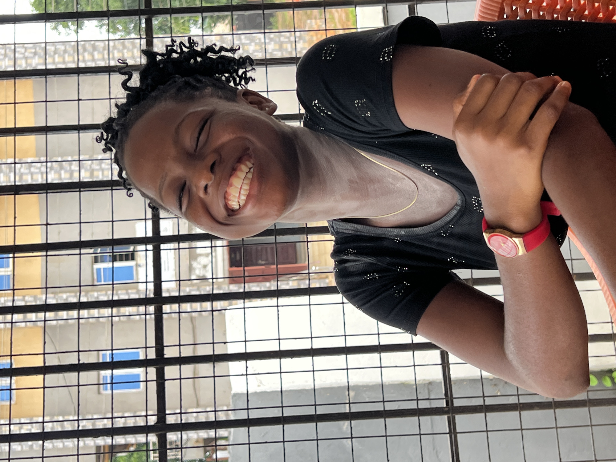 A young Black woman with curly hair and dark skin is shown smiling broadly with her eyes closed, wearing a black shirt and a red watch. She is seated in front of a black metal fence, which partially obscures a building with light-colored walls and blue windows. The environment suggests an outdoor setting, possibly in Nkpor, Nigeria, given the architectural style and vegetation visible in the background. The woman is leaning forward with her arms crossed, displaying a happy and relaxed demeanor.