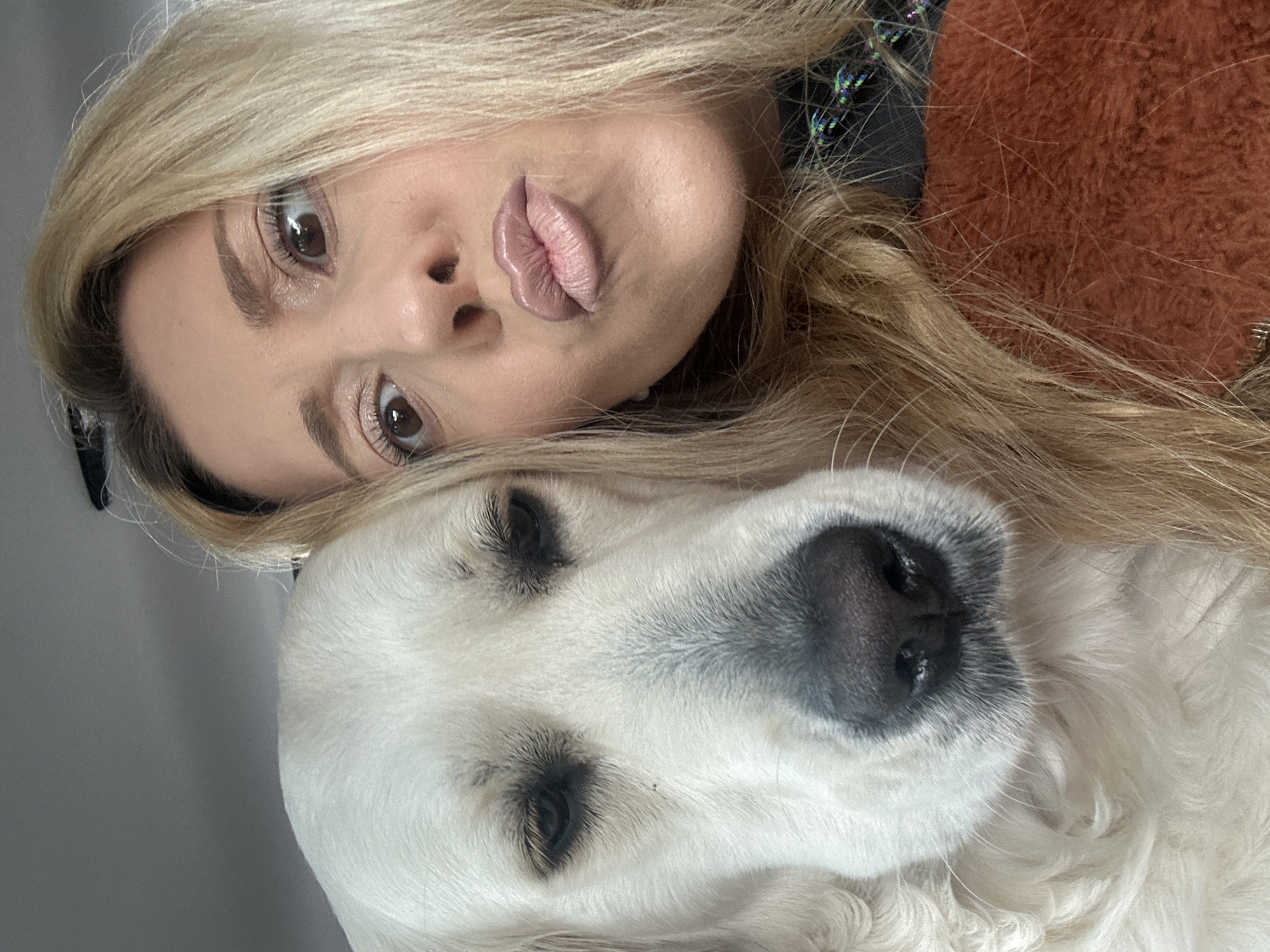 A woman with blonde hair is lying down, posing with her white dog. The woman's face is prominent, featuring pursed lips. Her hair is spread around her head. A white dog with black eyes and a black nose is lying next to her, with its head positioned beneath hers. The background is a plain gray. There is no indication of the location being in London, United Kingdom.
