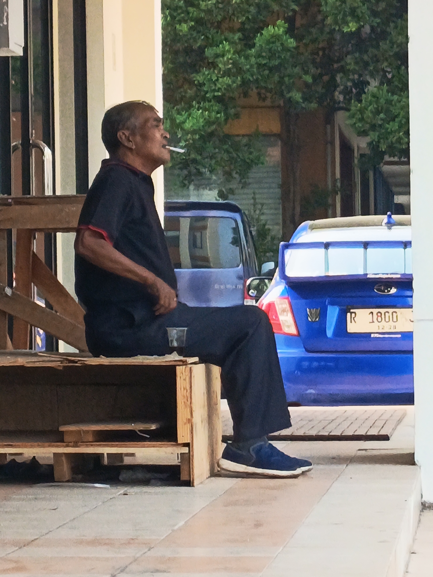 An elderly man with dark hair and a light complexion is sitting on a wooden bench outdoors. He is wearing a dark blue short-sleeved shirt with red trim on the collar and sleeves, and dark blue pants. A cigarette is lit and positioned in his mouth, and he is looking upwards and to his left. To his right, there is a small clear plastic cup on the bench. In the background, a blue Subaru WRX STI sedan is parked, with a license plate that reads "R 1800 12 28". Further in the background, a dark blue van is partially visible. The setting appears to be a public area with a tiled floor and some foliage in the background. The presence of the blue Subaru WRX STI with the specific license plate format suggests this scene is likely in Indonesia, as license plates with the "R" prefix are registered in the province of Banten, where Tangerang is located.