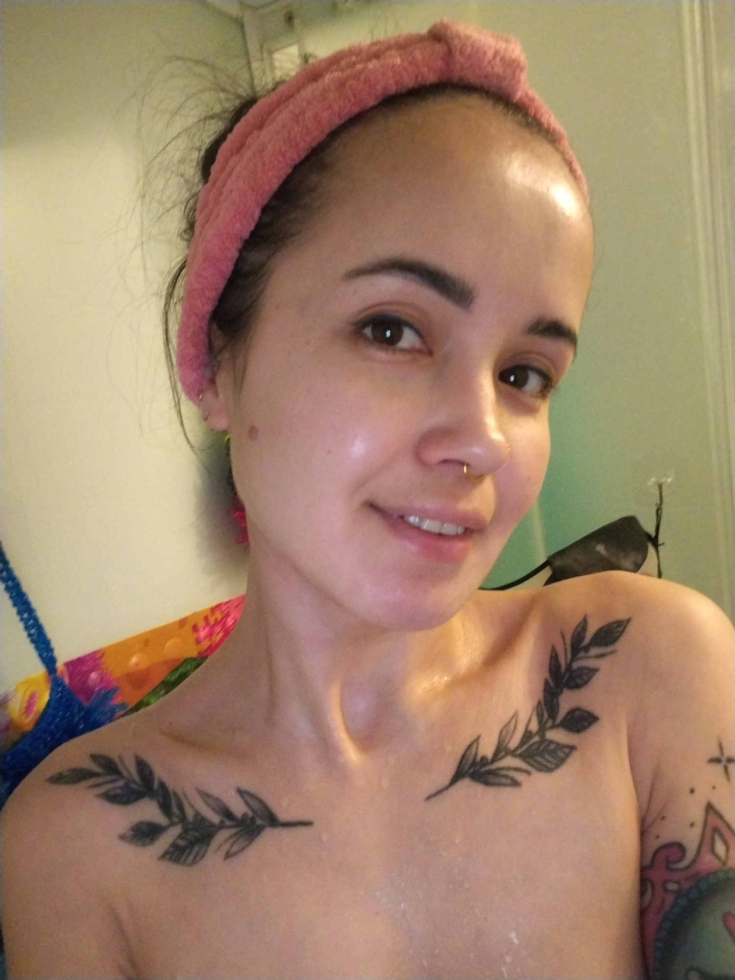 A person with light skin, dark hair pulled back with a pink headband, and a nose piercing is centered in the image. The person has dark eyebrows and brown eyes. The person is topless and has two tattoos of leafy branches across their upper chest, and additional tattoos on their left arm. The background appears to be a bathroom with a light green wall. A colorful mesh sponge is partially visible on the left side of the image. The person is smiling slightly and looking directly at the camera. There are droplets of water on the person's skin.
There is no indication of the location being in the city of Noviy, Russia.