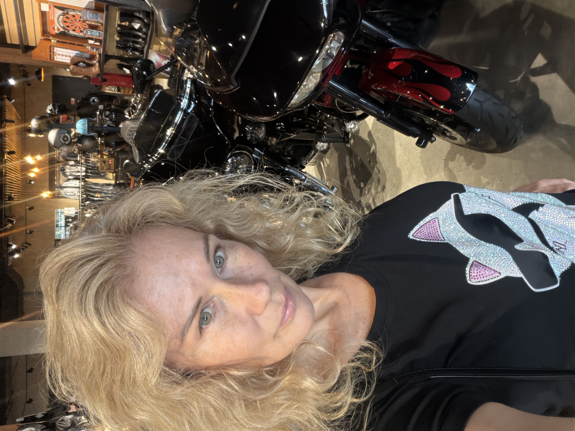 A fair-skinned woman with long blonde wavy hair is taking a selfie indoors. She is wearing a black t-shirt with a decorative design featuring pink and silver rhinestones. Behind her, a black motorcycle with red flame detailing is partially visible. The background shows a retail environment with various motorcycles and motorcycle accessories displayed, including helmets and dartboards. The location appears to be a motorcycle dealership. The city is not discernible from the image.