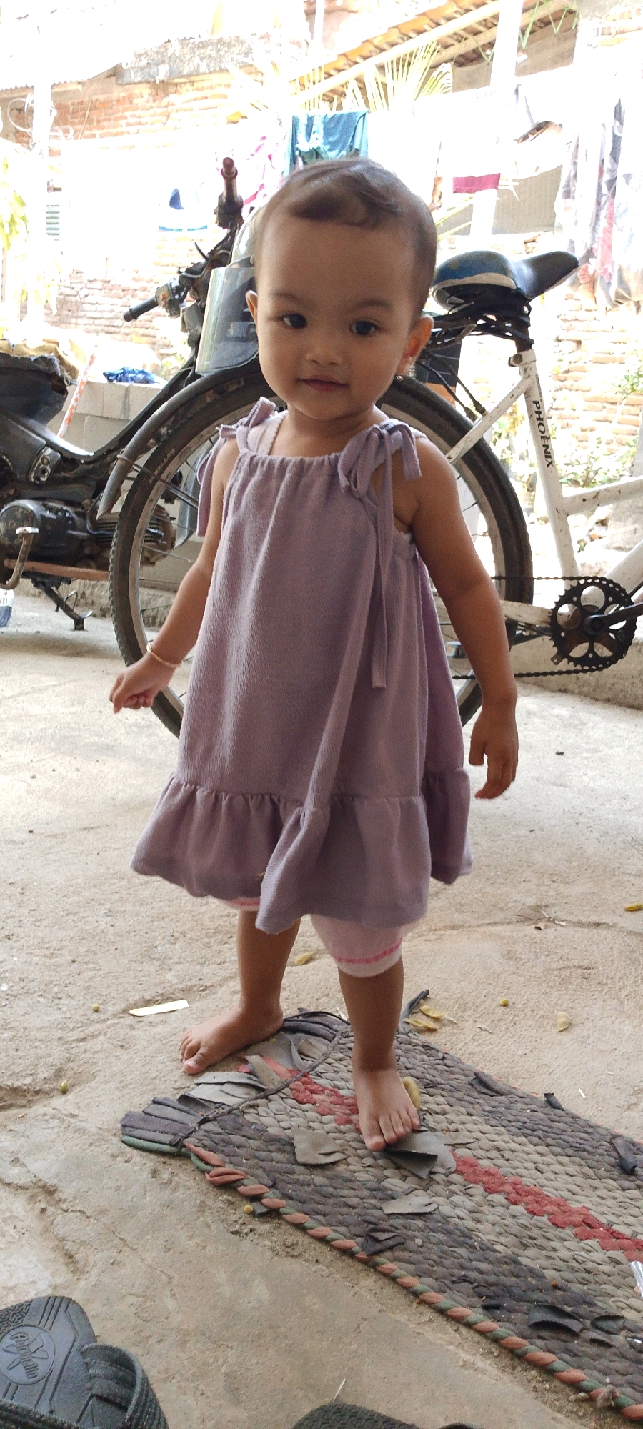 A young child, likely a toddler, stands barefoot on a woven mat. The child is wearing a light purple, ruffled dress over pink shorts. In the background, a white bicycle and a dark motorcycle are partially visible, along with a brick wall and laundry hanging on a line. The scene appears to be an outdoor residential area in Kamulyan, Indonesia, indicated by the architecture and typical rural setting. The ground is concrete, with a few scattered leaves and small pebbles.