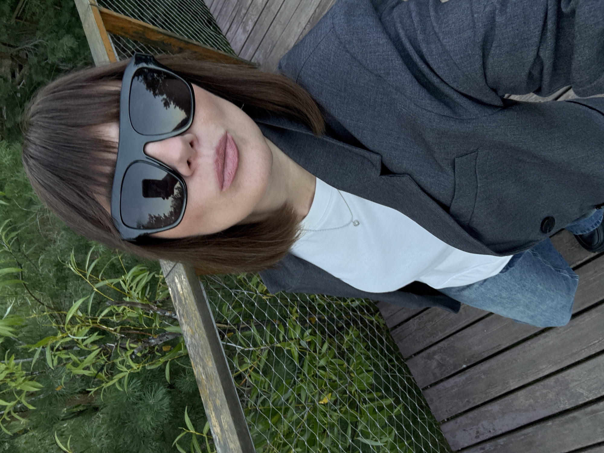 A woman with dark brown hair, bangs, and large black sunglasses stands on a wooden boardwalk surrounded by lush green foliage and trees. She is wearing a grey blazer over a white t-shirt and blue jeans. The boardwalk has a wooden railing and a wire mesh fence. The reflection in her sunglasses shows a smartphone being used to take the selfie and trees. The scene appears to be in a park or nature area, possibly the bor, Russia.