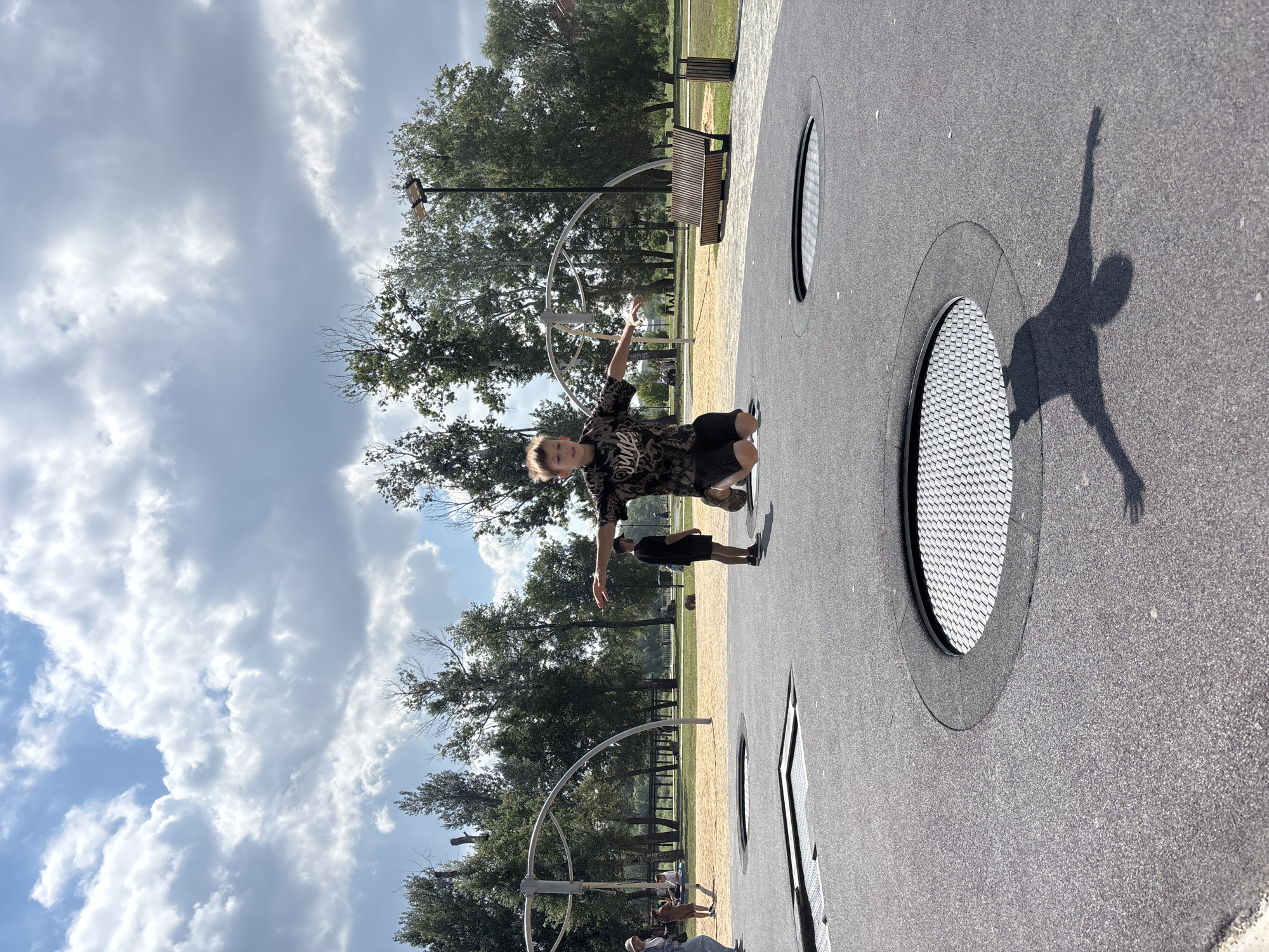 In Orenburg, Russia, a young boy is captured mid-air, jumping on an in-ground trampoline in a park. He is wearing a black t-shirt with a graphic print and black shorts, with his arms outstretched. His shadow is visible on the trampoline surface, mimicking his airborne pose. The park features several other in-ground trampolines, some of which are also visible in the frame. In the background, trees and a cloudy sky create a natural setting. Other people are present in the park, including adults and children, some of whom are walking and others are seated. A wooden bench and trash receptacle are also visible.