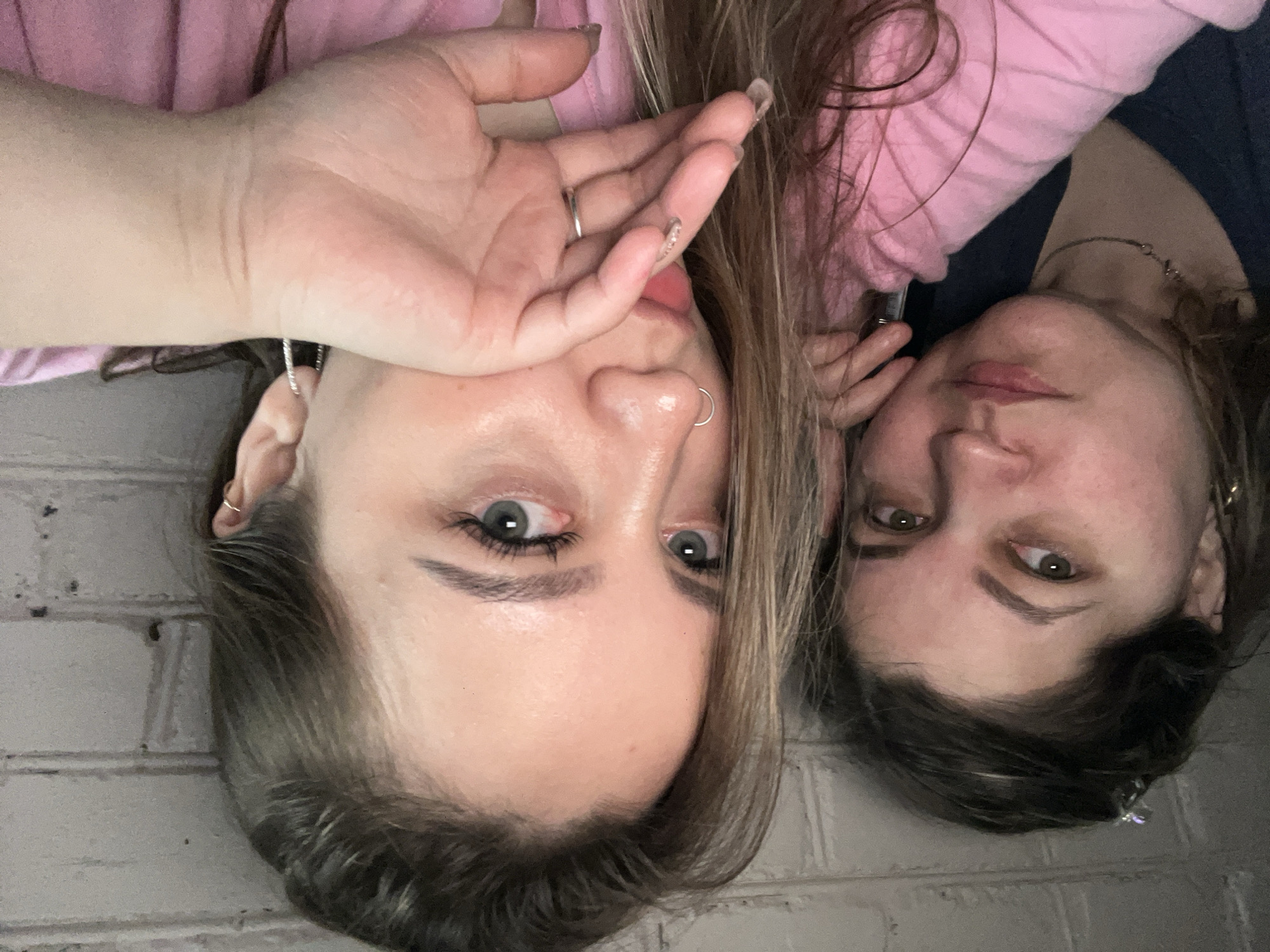 Two Caucasian women are closely positioned side-by-side. The woman on the left has light skin, light brown hair, blue eyes, a nose ring, and several earrings. Her right hand is raised to her face, partially covering her mouth. She wears a pink top. The woman on the right has light skin, dark brown hair, and green eyes. She wears a dark-colored top and a necklace. The background appears to be a light-colored brick wall. The image appears to have been taken with the camera rotated 180 degrees. The image provides no information connecting it to Novopokrovskaya, Russia.