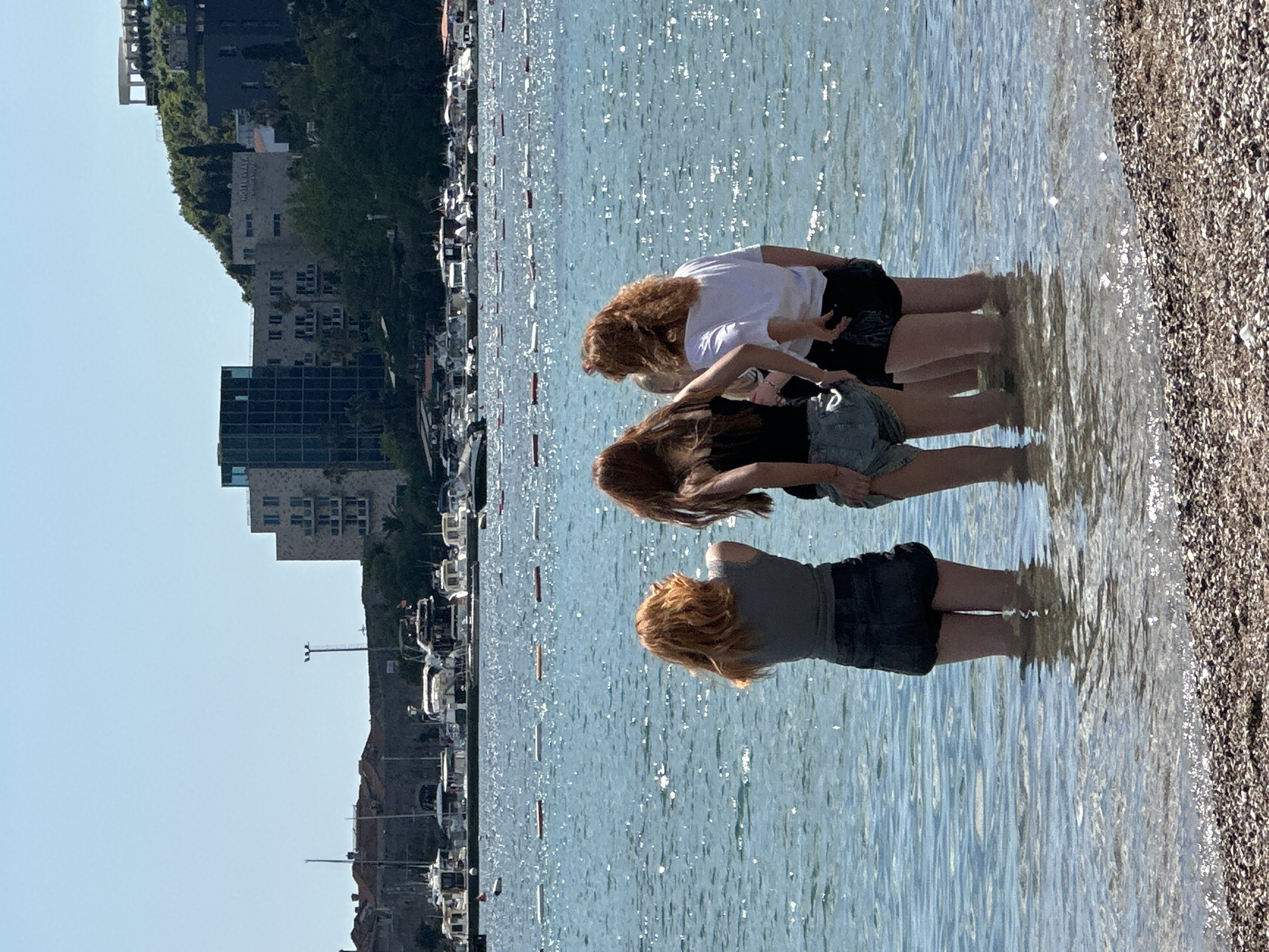 Three individuals are standing in shallow water along a pebbled beach, likely in Budva, Montenegro, given the presence of buildings, possibly a marina or harbor, and mountainous terrain in the background. The person closest to the viewer has auburn hair, is wearing a gray shirt and dark-colored shorts. The middle person has long brown hair and is wearing a black shirt and lighter-colored shorts. The furthest person has auburn hair and is wearing a white shirt and dark-colored shorts. All individuals appear to be standing, facing toward the water with their legs submerged. The water is light blue and reflective, and the sky is clear. Boats are moored in the distance.