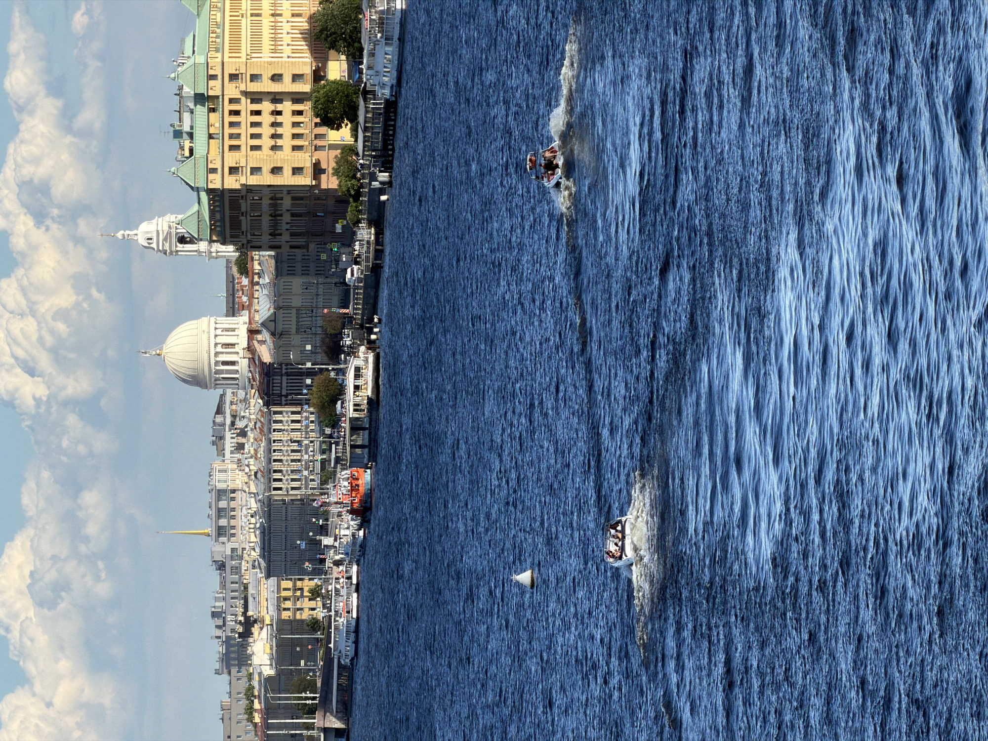 This is a wide-angle, medium-distance photograph of the Neva River in Saint Petersburg, Russia, on a clear, sunny day with some clouds in the sky. The viewpoint is elevated, looking across the river towards the city's historic architecture.
In the foreground, the deep blue water of the Neva River is textured with moderate waves. Two small motorboats are navigating the river. The boat closer to the viewer and slightly to the left has several passengers visible, its wake trailing behind. Further away and to the right, another motorboat with passengers is also moving, creating a white wake. A solitary white buoy is visible in the water between the two boats.
The middle ground of the image showcases the St. Petersburg skyline. Along the riverbank, numerous passenger boats are docked. These include several white tour boats and a prominent orange and white vessel in the center. The embankment is lined with buildings featuring classical architectural styles, indicative of the city's historical significance. Prominent landmarks visible include a large, white domed building (likely St. Isaac's Cathedral) and a slender, golden spire (likely the spire of the Peter and Paul Fortress). Other buildings include a distinctive yellow building with a green roof on the right side of the frame, and various other multi-story structures with ornate facades. Trees are interspersed along the embankment.
The background consists of a bright blue sky with fluffy white clouds. The overall impression is a vibrant and active river scene against the backdrop of a grand European city.