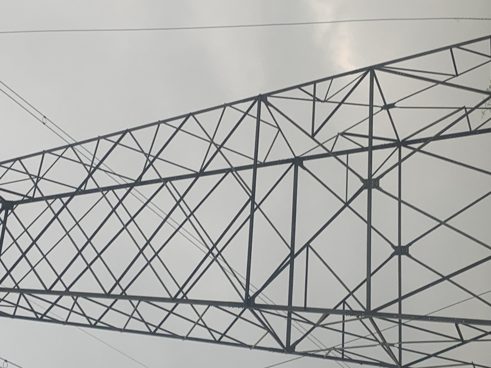 The image shows a close-up, low-angle view of a tall, latticed metal power transmission tower against a cloudy sky. The tower is composed of dark metal beams forming a geometric, triangular structure that tapers towards the top. Several electrical wires are visible, running diagonally across the frame and horizontally from the tower. The overall lighting is diffused due to the overcast conditions. The scene appears to be outdoors, in an area where such infrastructure is present, but no specific urban landmarks or features are discernable.