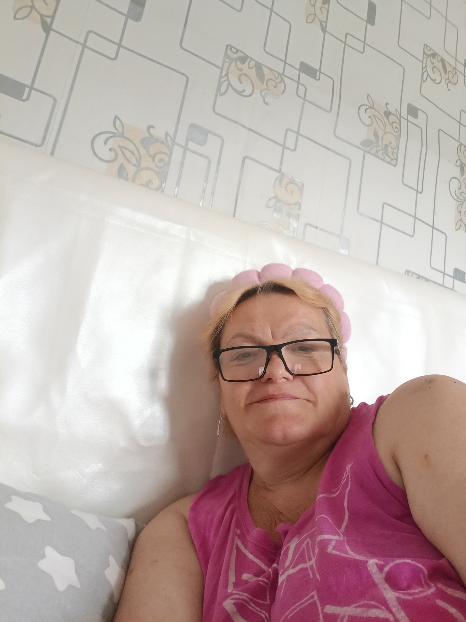 A woman with blond hair and black eyeglasses is lying down. She is wearing a pink, puffy headband and a pink sleeveless shirt. She is propped up against a white padded headboard. To her left is a grey pillow with white stars. The wall behind the headboard is white with a geometric pattern. The image does not provide enough information to confirm the location is Красный Маяк, Russia.