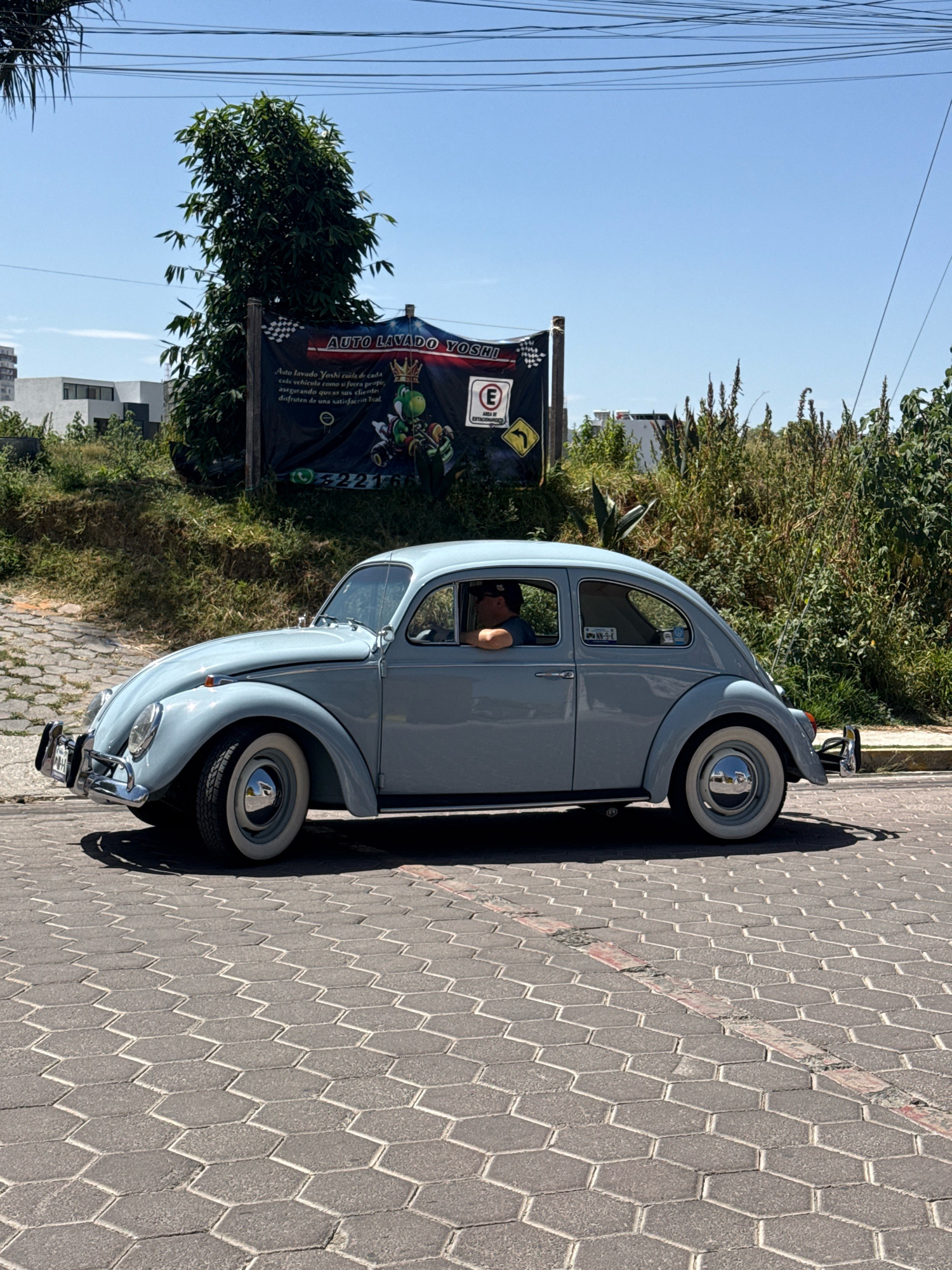 A light blue vintage Volkswagen Beetle is parked on a hexagonal paver street in Santa Clara Ocoyucan, Mexico. The car is facing left and is slightly angled towards the viewer. A person wearing a dark t-shirt and a baseball cap is visible in the driver's seat, resting their arm on the open window. The car has whitewall tires with chrome hubcaps. Behind the car, a black banner hangs from a wooden post, advertising "AUTO LAVADO YOSHI." The banner features an image of the cartoon character Yoshi. A "P" sign indicating parking is also visible on the banner, along with a yellow directional sign. Green foliage and trees are behind the banner and to the right of the car. Buildings can be seen in the background. The sky is clear and blue, indicating a sunny day.