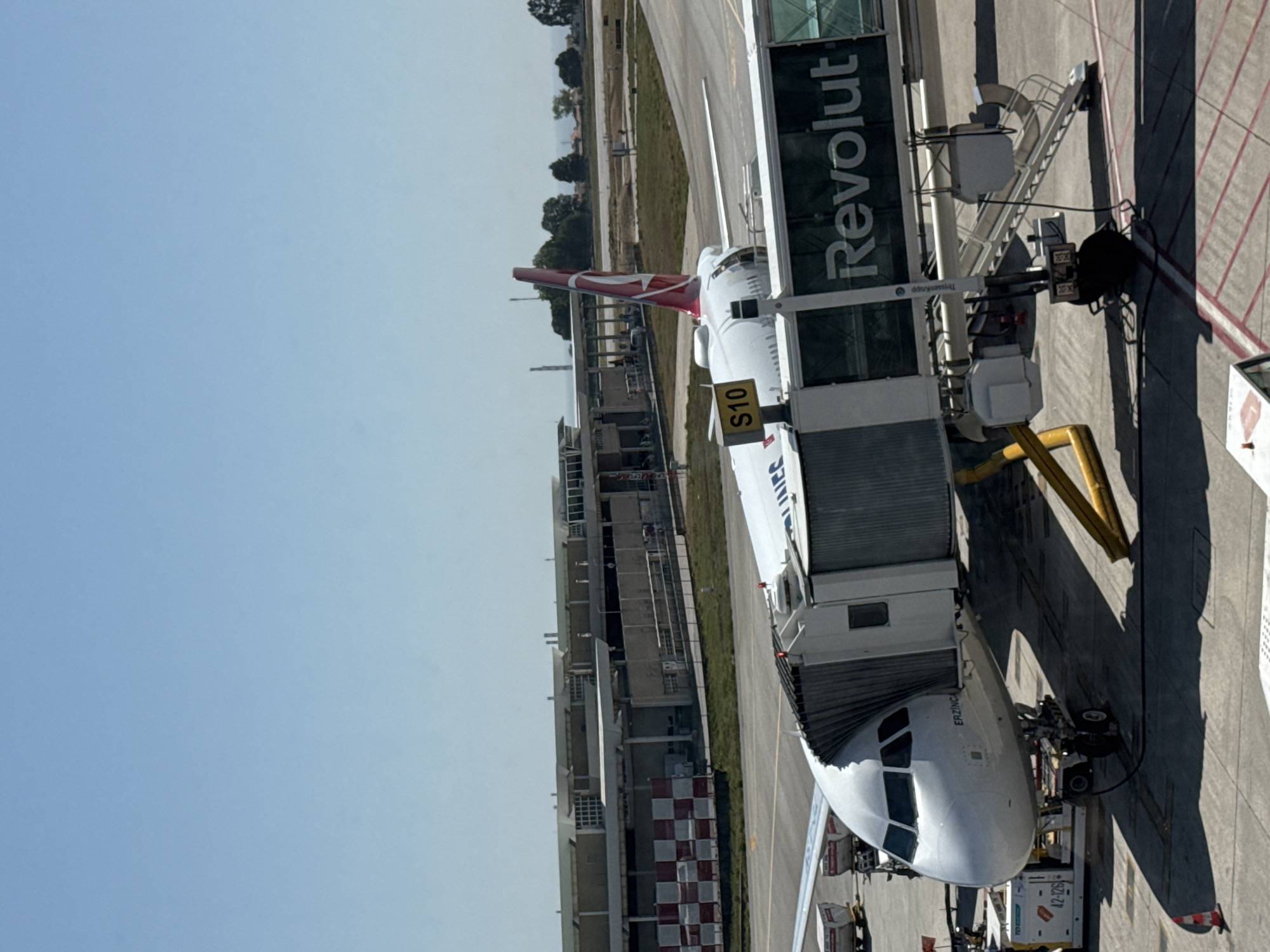 This is a medium-shot, eye-level, outdoor, daytime photograph of an airplane at an airport gate. The airplane, a Turkish Airlines Airbus A320 with the registration TC-JRY and the name "ERZINCAN" visible on its fuselage, is connected to a jet bridge. The jet bridge is labeled "Revolut" and has a yellow sign with "S10" on it. In the background, a terminal building of Porto Airport (OPO) in Matosinhos, Portugal, can be seen. The airport tarmac is visible, as are various ground support equipment, including baggage carts and a tow tractor. The sky is clear and blue. There are no people clearly visible in the image. The overall scene depicts a typical airport operation with an aircraft at the gate.