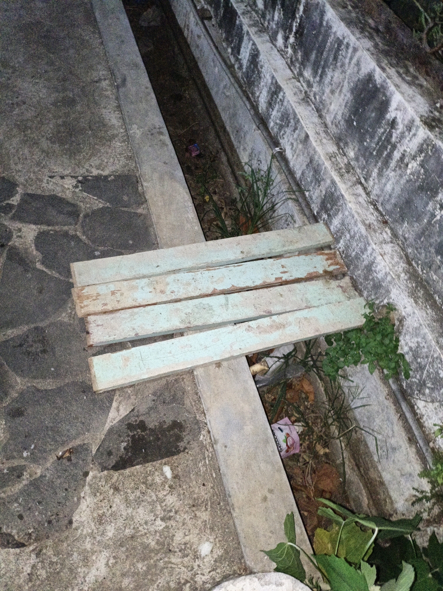 In Cikal, Indonesia, a temporary bridge is constructed over a drainage ditch using four weathered, light blue wooden planks. The planks are positioned horizontally, spanning the ditch, with the left side resting on a rough, dark grey stone pavement and the right side supported by a grey concrete curb. The drainage ditch below is filled with dry leaves, sparse green vegetation, and a small piece of pink and white wrapper. To the left of the ditch, a section of dark grey stone pavement is visible, with a pattern of irregularly shaped stones. To the right, a textured, grey concrete wall with vertical divisions and patches of white and black discoloration runs parallel to the ditch. Green foliage is present at the bottom right of the image, encroaching into the scene. The lighting suggests it is either dusk or night.