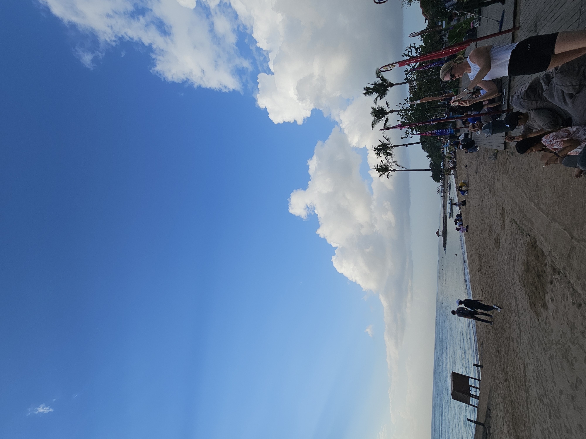 This image depicts a scene at a beach in Denpasar, Indonesia, likely Kuta Beach, given the prominent palm trees and beachside structures often found there. The sky is a vibrant blue with large, fluffy white clouds scattered across it. In the foreground, a sandy beach stretches towards the ocean, with the calm, blue water meeting the shore.
Several people are present on the beach and a wooden boardwalk area. On the beach, two individuals are walking near the water's edge. Further back, groups of people are seated on beanbag chairs or benches, some engaged with their phones. A woman in a white top and black shorts is prominently featured in the mid-right, holding a phone and appearing to take a picture. Behind her, several tall, thin flagpoles with red and white banners are visible, along with several palm trees lining the background.
To the left, a wooden structure on stilts stands near the water, and a few boats are visible in the distance. In the far distance, a small gazebo-like structure can be seen on the horizon. The overall atmosphere suggests a relaxed, tourist-oriented beach environment during the day.