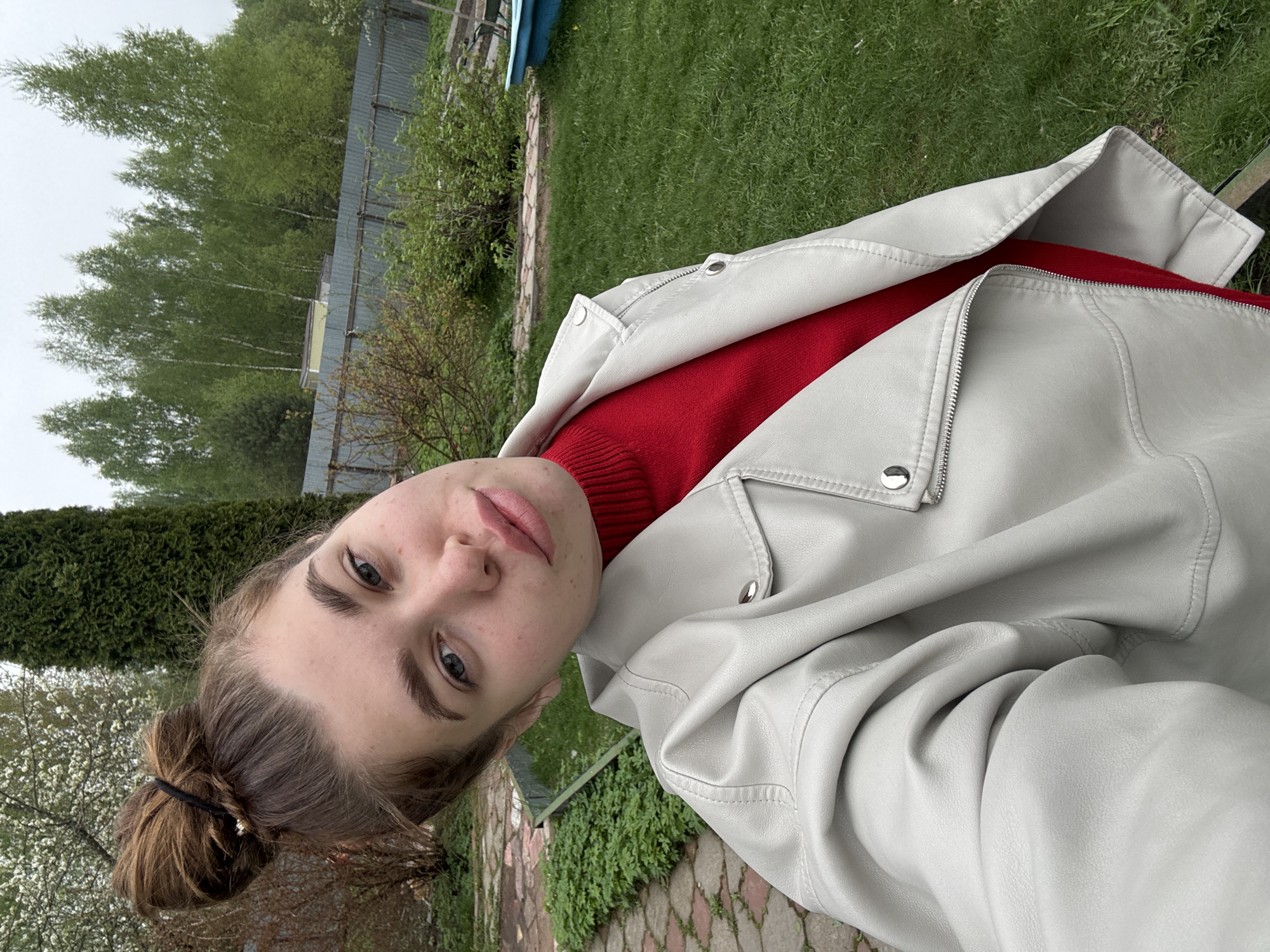 A woman is standing in Мещёры, Russia, likely in a garden or outdoor area. She is wearing a red turtleneck sweater and a light-colored leather jacket. Her hair is pulled back in a bun. The background includes trees, a fence, and greenery.
