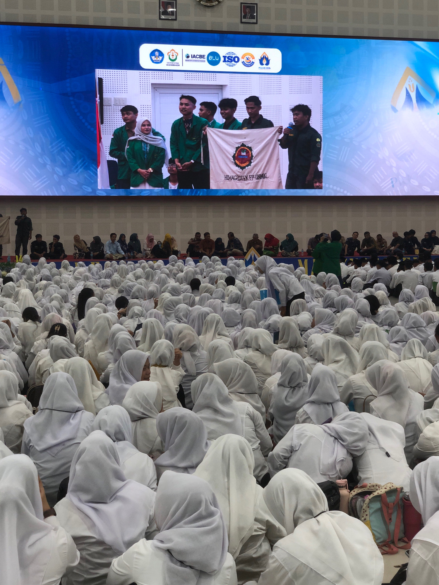 The image depicts a large gathering of people, primarily young individuals wearing white attire, seated on the floor in an indoor venue. The audience is densely packed, with many individuals wearing white hijabs. In the background, a large screen displays a presentation with logos and images of people. A group of individuals are on a stage in front of the screen, some holding a banner that reads "HIMAGROTEK FP-UNIMAL." The audience is looking towards the stage. The event appears to be a formal gathering or ceremony, possibly at a university or educational institution. The location in Reuleut Timu, Indonesia is indicated by the text on the banner.