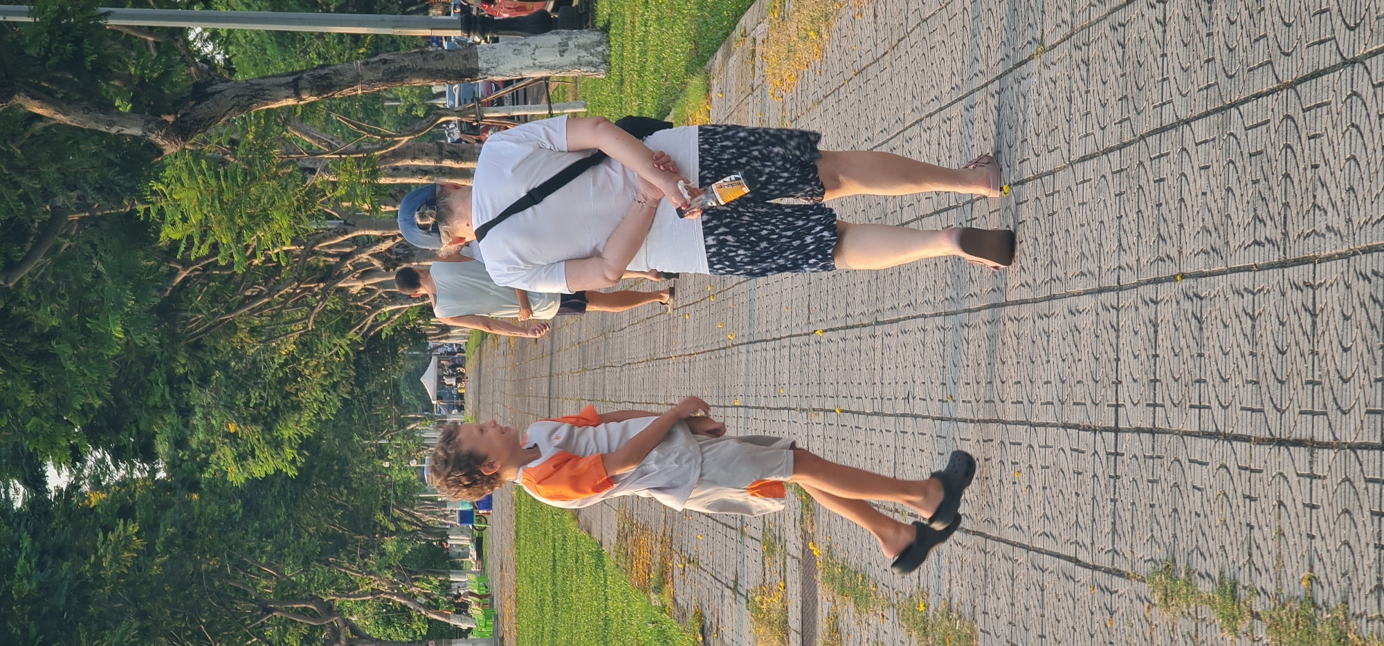 The image captures a vibrant late afternoon scene in what appears to be a public park or promenade in Đà Nẵng, Vietnam. The sun casts warm light and distinct shadows, suggesting either morning or late afternoon.
In the foreground, a young boy with curly hair, wearing an orange and white short-sleeved shirt, light shorts, and black clogs, stands on a patterned paved path. He is looking upwards and slightly to his right, with his mouth open, seemingly talking or laughing.
Behind him, an adult person, viewed from the back, walks along the path. This individual wears a white t-shirt, black and white patterned shorts, and light-colored flat shoes, along with a blue baseball cap and a black cross-body bag. They are holding a small brown bottle with an orange label behind their back with both hands.
Further down the path, another adult in a light-colored t-shirt and dark shorts is also walking. In the distance, several indistinct figures and what appear to be market stalls or tents are visible, hinting at a lively public space.
The wide pathway is made of gray pavers with a distinct repeating geometric pattern. Scattered yellow petals or leaves dot the path, adding a touch of color. Lush green grass borders the path on both sides, and dense tropical trees with prominent roots and branches line the area, creating a leafy canopy. A metal pole and some parked vehicles are partially visible in the upper background. No clear legible text is visible on any objects in the image.