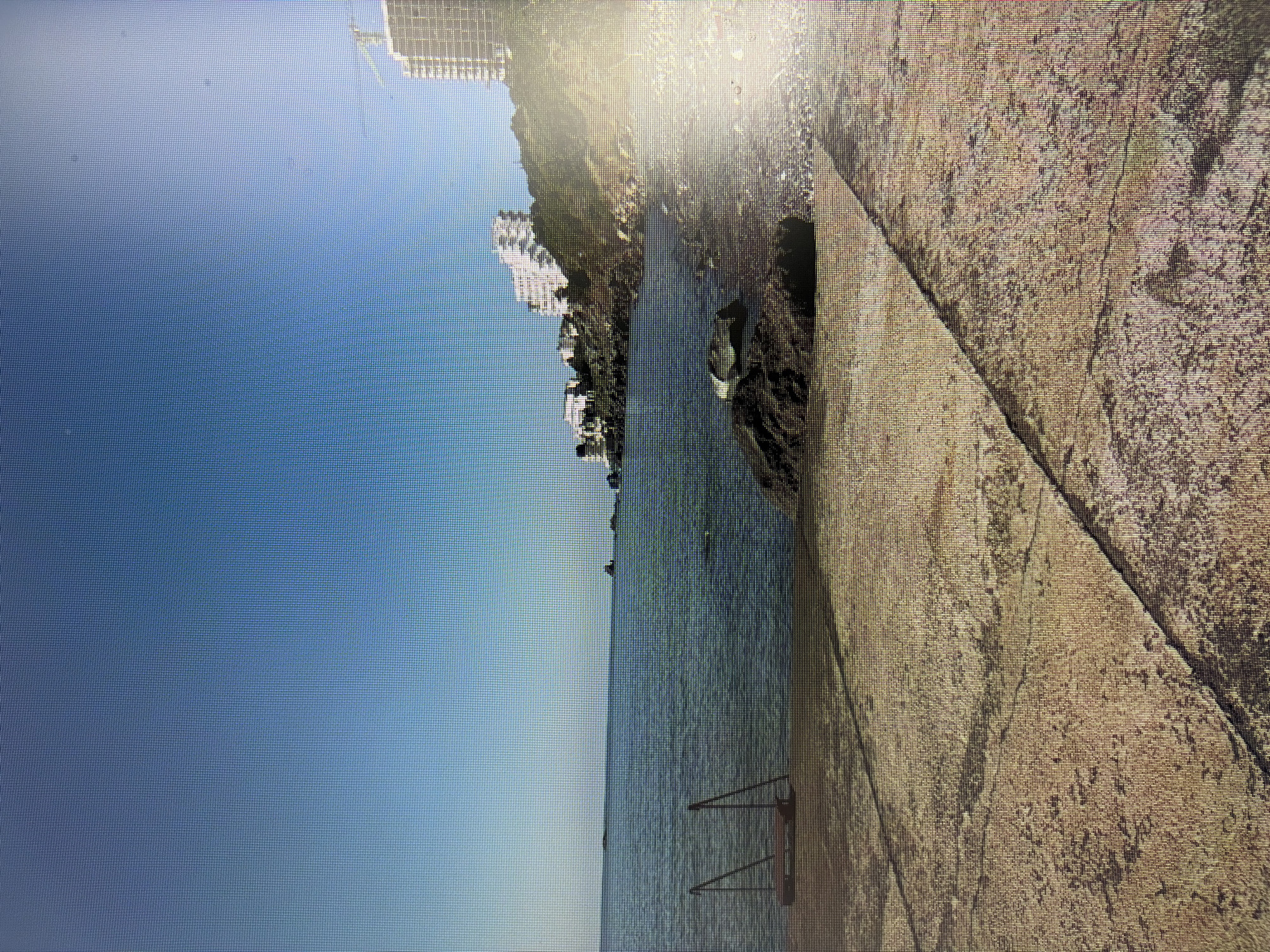The image depicts a waterfront scene with a concrete walkway in the foreground. To the left of the walkway, a set of metal steps leads into the calm, blue water. The water extends to the horizon, meeting a clear blue sky. Along the coastline in the distance, several white buildings are visible on a hillside, with a construction crane towering over one of them. The environment suggests a sunny day at a coastal location. The image does not provide any information about the city of Mirnoye, Ukraine, or any specific details about people or actions.