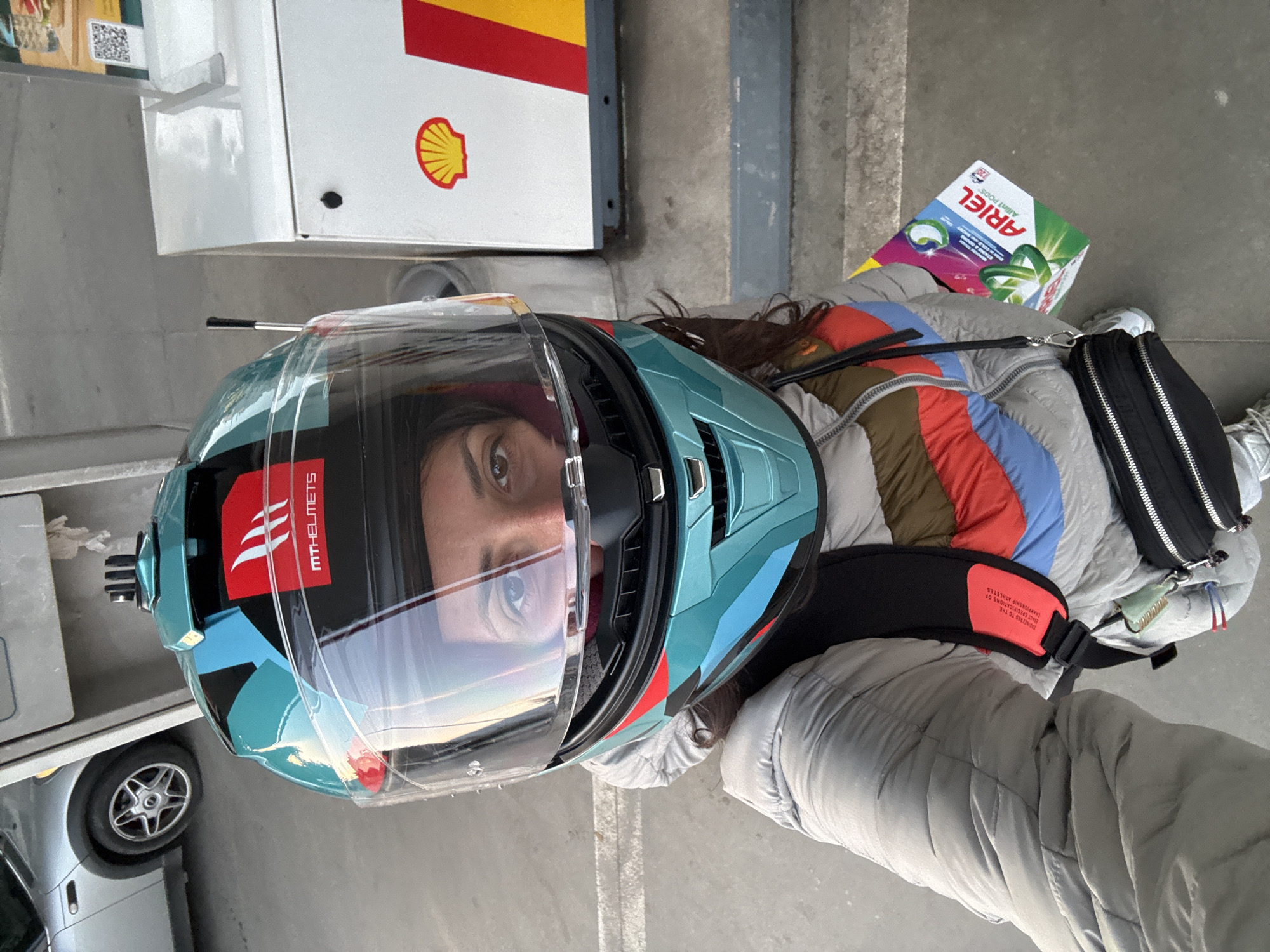 A woman wearing a blue motorcycle helmet with a transparent visor is standing at a Shell gas station. She is wearing a puffy gray jacket with colorful stripes, and a black shoulder bag. She is holding a box of Ariel laundry pods. A silver car is visible in the background. The scene is located in Sofia, Bulgaria.