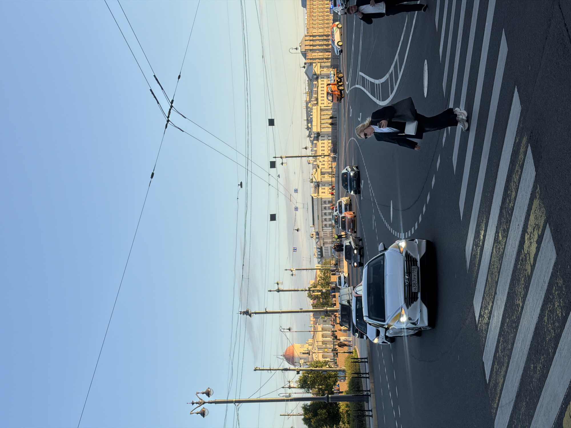 This image captures a street scene in Saint Petersburg, Russia, likely near the Palace Square or Admiralty Embankment, given the architectural style of the buildings in the background. The sky is clear and a pale blue, indicating it is either early morning or late afternoon. Overhead, trolleybus wires are visible, crisscrossing the frame.
In the foreground, a zebra crossing is prominently featured on the asphalt road. A woman in a black blazer and pants, carrying a large gray tote bag, is walking across the zebra crossing from left to right. She is wearing sunglasses and white sneakers.
Several cars are present on the road. A white sedan, possibly a taxi, with yellow accents and a license plate reading "H387PY 198", is stopped at the crossing. Other vehicles, including a dark sedan and an orange truck, are visible further down the road.
In the background, grand classical buildings with ornate facades and columns line the street. One building features a prominent red dome. People are seen walking on the sidewalks and near the buildings. Streetlights and decorative lampposts are spaced along the thoroughfare. The overall impression is that of a busy urban environment bathed in the soft light of the setting or rising sun.