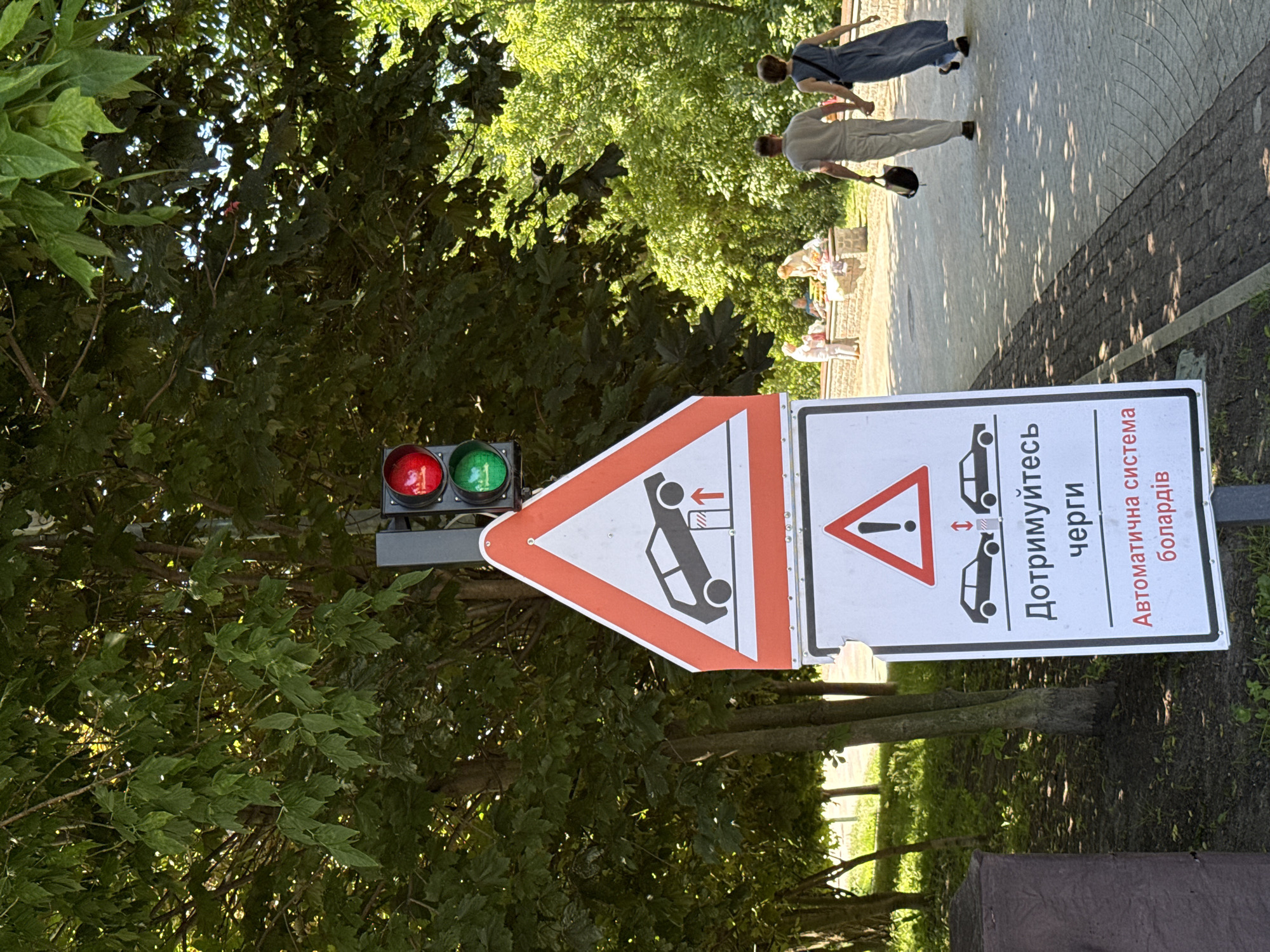 This is a medium shot of a traffic light and two signs in Kyiv, Ukraine. The traffic light is red on top and green on the bottom, indicating that traffic is currently allowed to proceed. The first sign, a triangular warning sign with a red border, depicts a car being lifted by a hydraulic jack. Below it, a rectangular sign with a red border displays an exclamation mark within a triangle, a common symbol for a warning. Underneath the exclamation mark, there are two black car silhouettes with a red double-headed arrow between them. Below the car graphics, text reads "Дотримуйтетесь черги" (which translates to "Observe the queue") followed by "Автоматична система болардів" (which translates to "Automatic bollard system"). The signs are positioned on a paved walkway bordered by greenery and trees. In the background, two people, a man and a woman, are walking away from the camera on the walkway, holding hands. Further in the background, several other individuals are seated near a low stone structure. The scene appears to be a sunny day, with dappled light filtering through the trees onto the walkway.