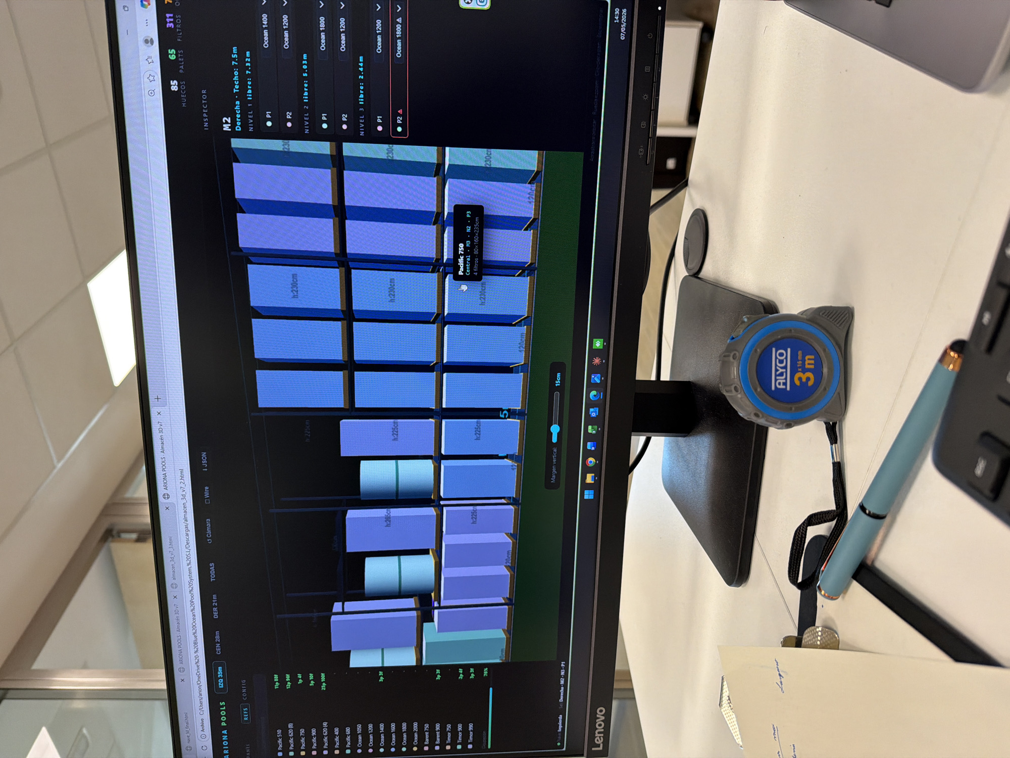 The image shows a desk with a Lenovo monitor displaying a 3D rendering of a warehouse layout with shelves and inventory. The screen shows a browser window with a software application called "Ariona Pools Almacen 3D v7" open. On the right of the monitor, there is a blue and gray "ALYCO" branded 3-meter measuring tape, a light blue pen, a keyboard, and a small piece of paper with writing on it. The desk appears to be in an office environment. The software interface indicates that the warehouse design is being analyzed, showing dimensions and specifications for each section and item within the warehouse. The time displayed in the corner of the screen is 14:30 on 07/05/2026. The software shows details of "Ocean" and "Pacific" brand inventory along with dimensions in centimeters. There is no clear indication of Errenteria, Spain.